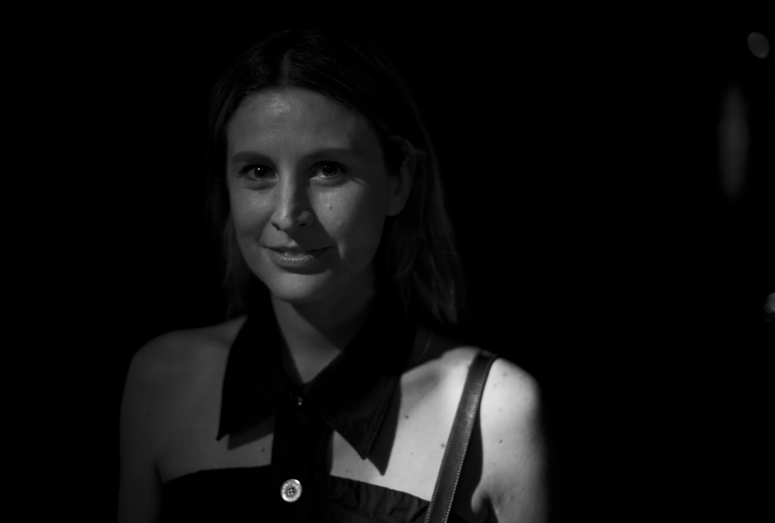 Portrait en noir et blanc d'une femme avec une expression douce, portant une tenue noire avec un col en forme de nœud, dans un environnement sombre.