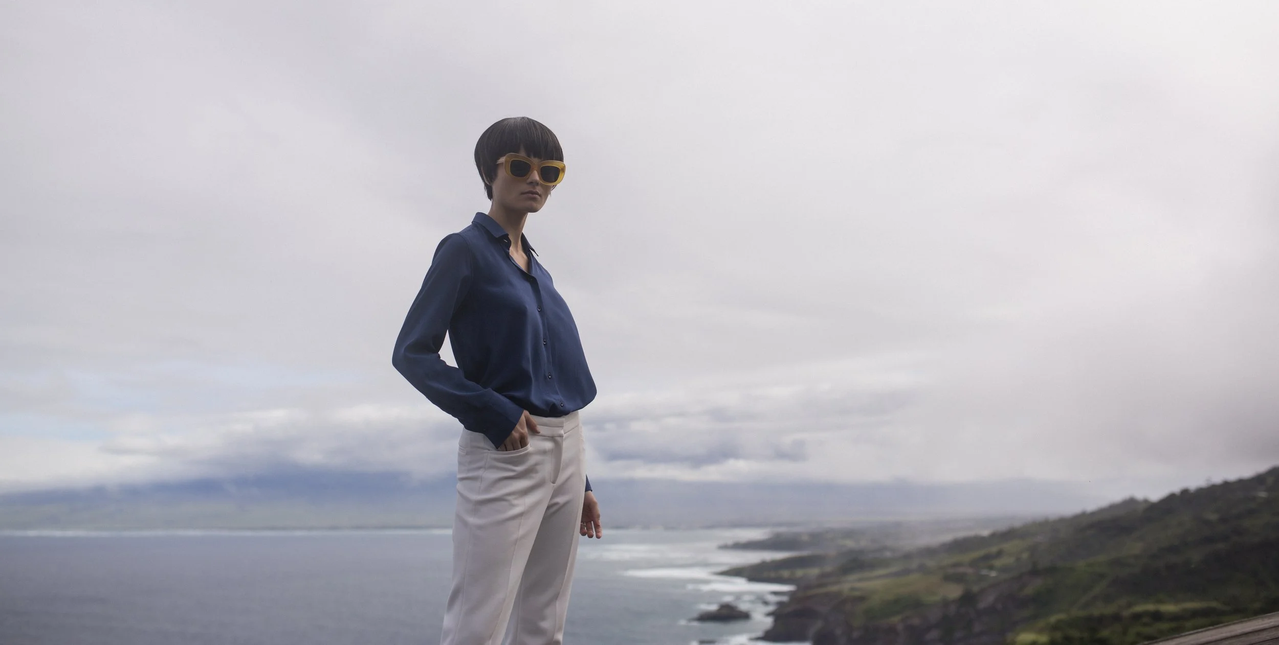 Une femme portant des lunettes jaunes, une chemise bleue et un pantalon beige se tient sur un promontoire avec une vue sur la côte et la mer sous un ciel nuageux.