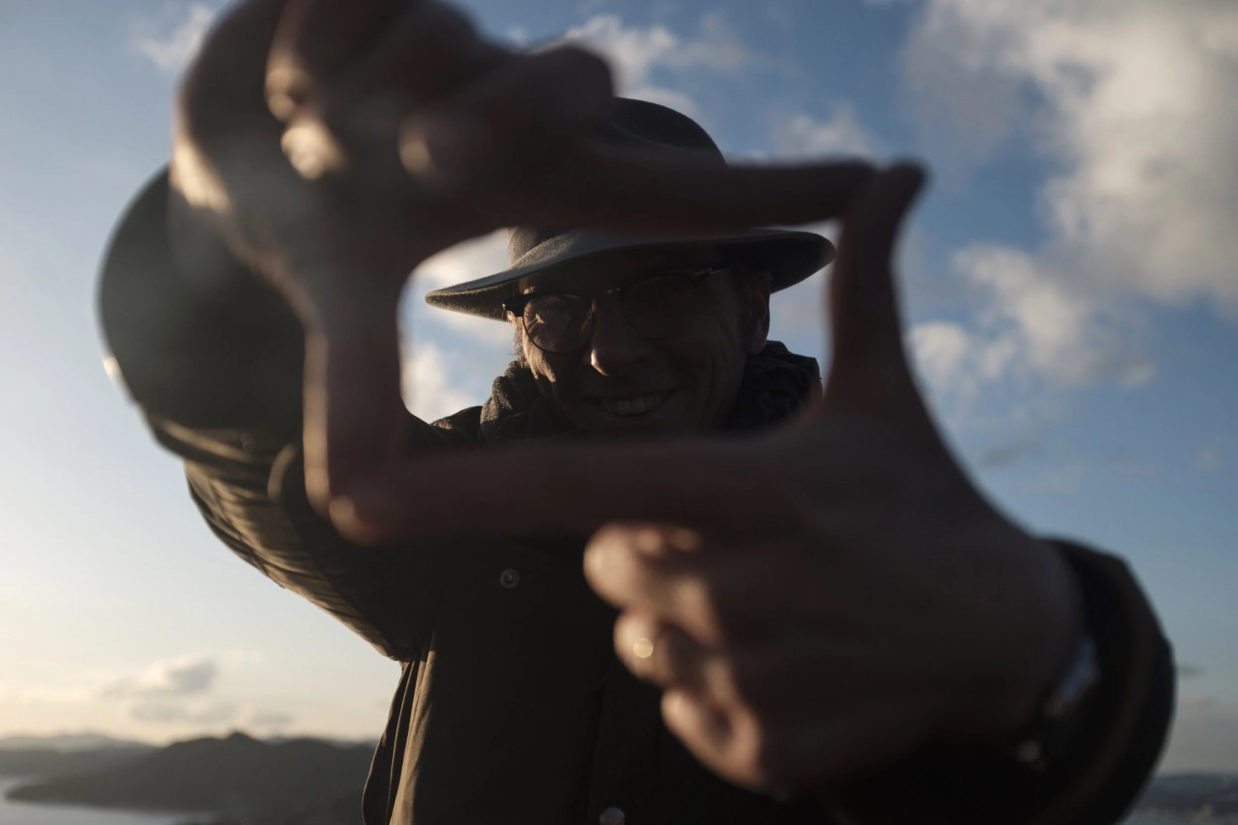 Une personne portant un chapeau et des lunettes regarde à travers un cadre, avec un ciel partiellement nuageux et une montagne en arrière-plan.