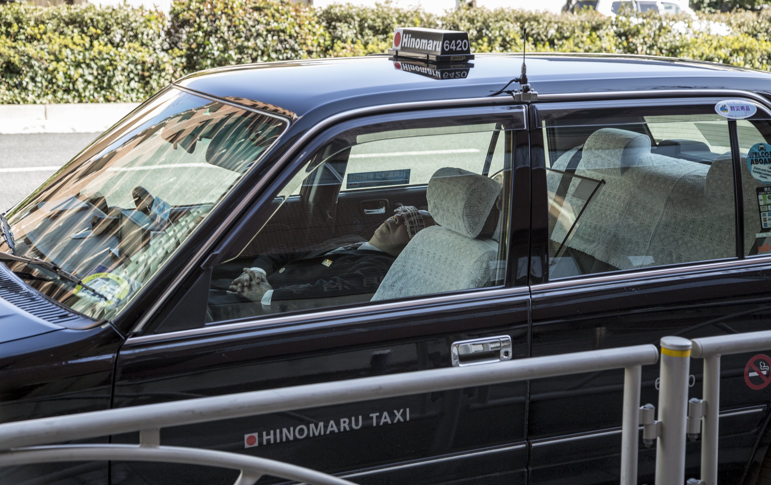 Un taxi noir avec des sièges recouverts de housses en dentelle, une personne allongée à l'intérieur, vue à travers la fenêtre, avec des panneaux de signalisation et une haie en arrière-plan.