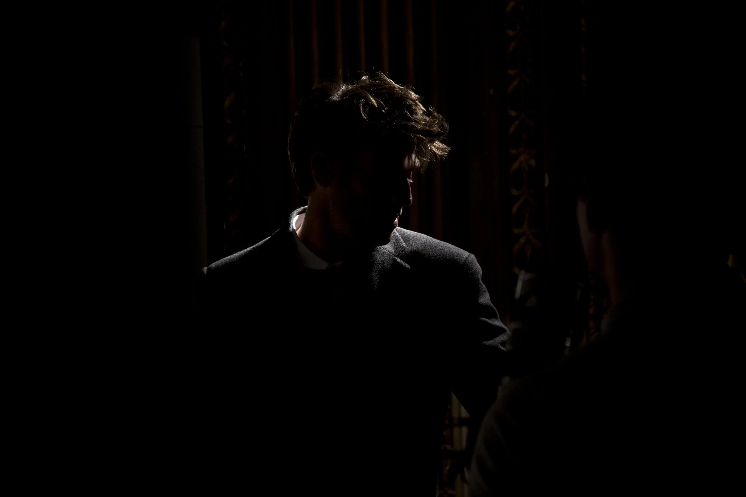 Silhouette d'un homme avec cheveux bouclés, portant un costume, dans une pièce sombre avec un éclairage venant de derrière.