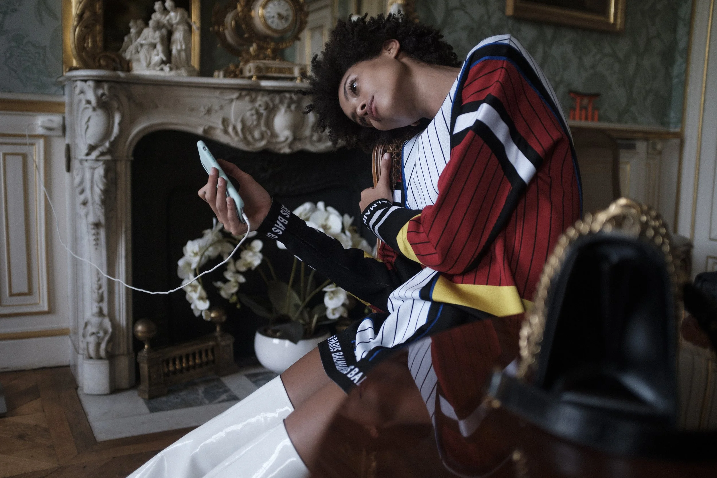Une femme assise sur une chaise en ornement et en tissu noir, portant une robe multicolore, regarde un téléphone portable connecté par câble USB, dans une pièce décorée avec un cheminée en marbre, un miroir doré, et des fleurs blanches dans un vase b