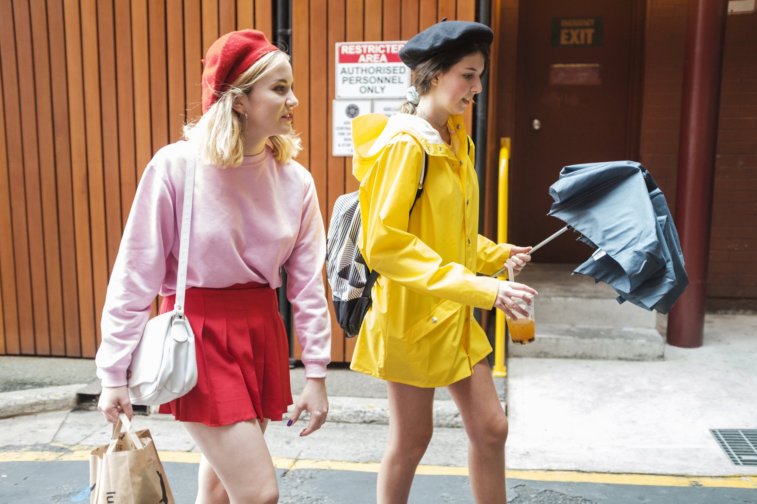 Deux jeunes femmes marchant dans la rue, l'une porte un sweat rose, une jupe rouge et un sac blanc, l'autre une veste jaune, tenant un parapluie et une boisson, en arrière-plan un mur en bois et des panneaux de signalisation.