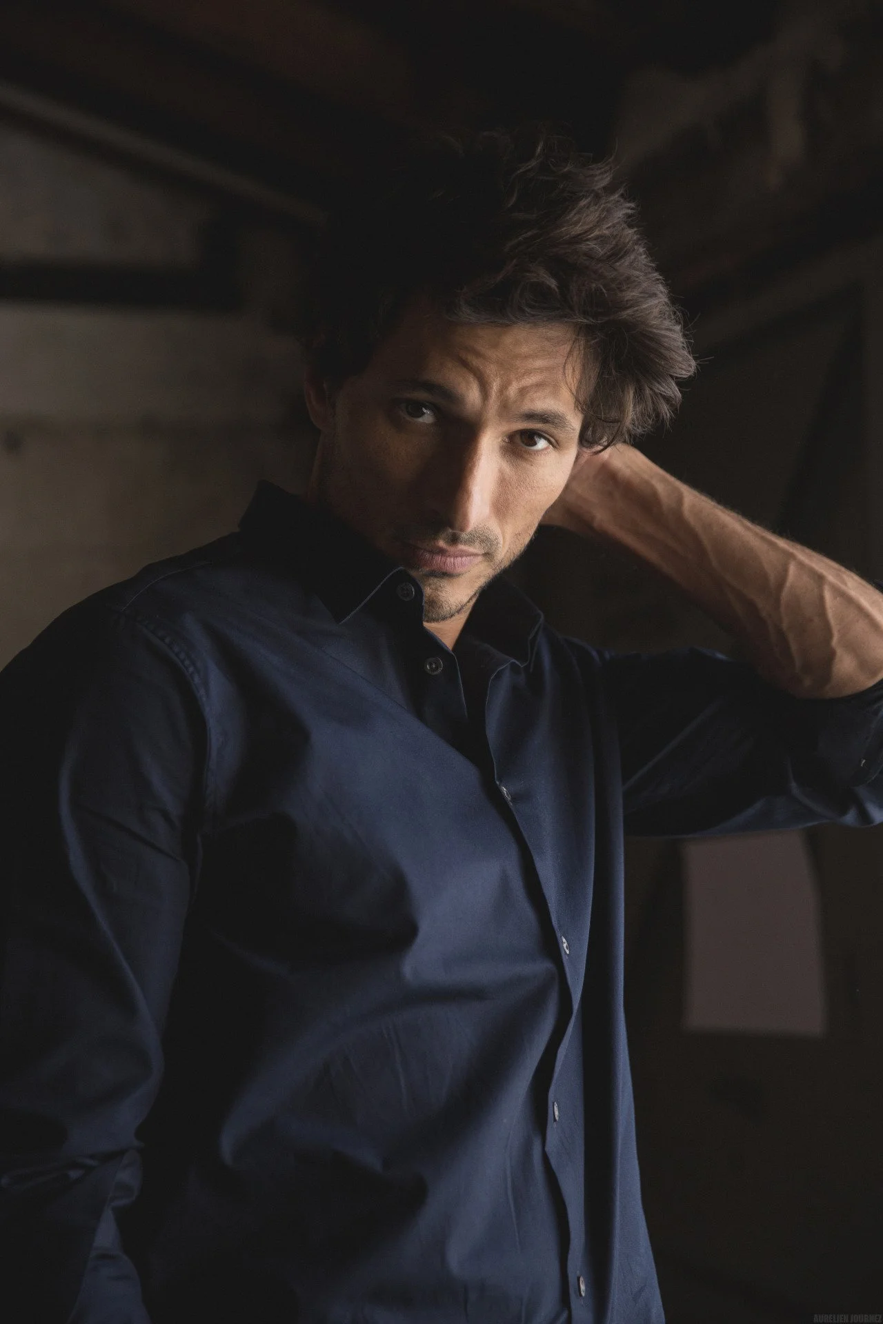 Un homme portant une chemise bleu foncé, posé dans un environnement sombre, avec une expression sérieuse et regardant la caméra.