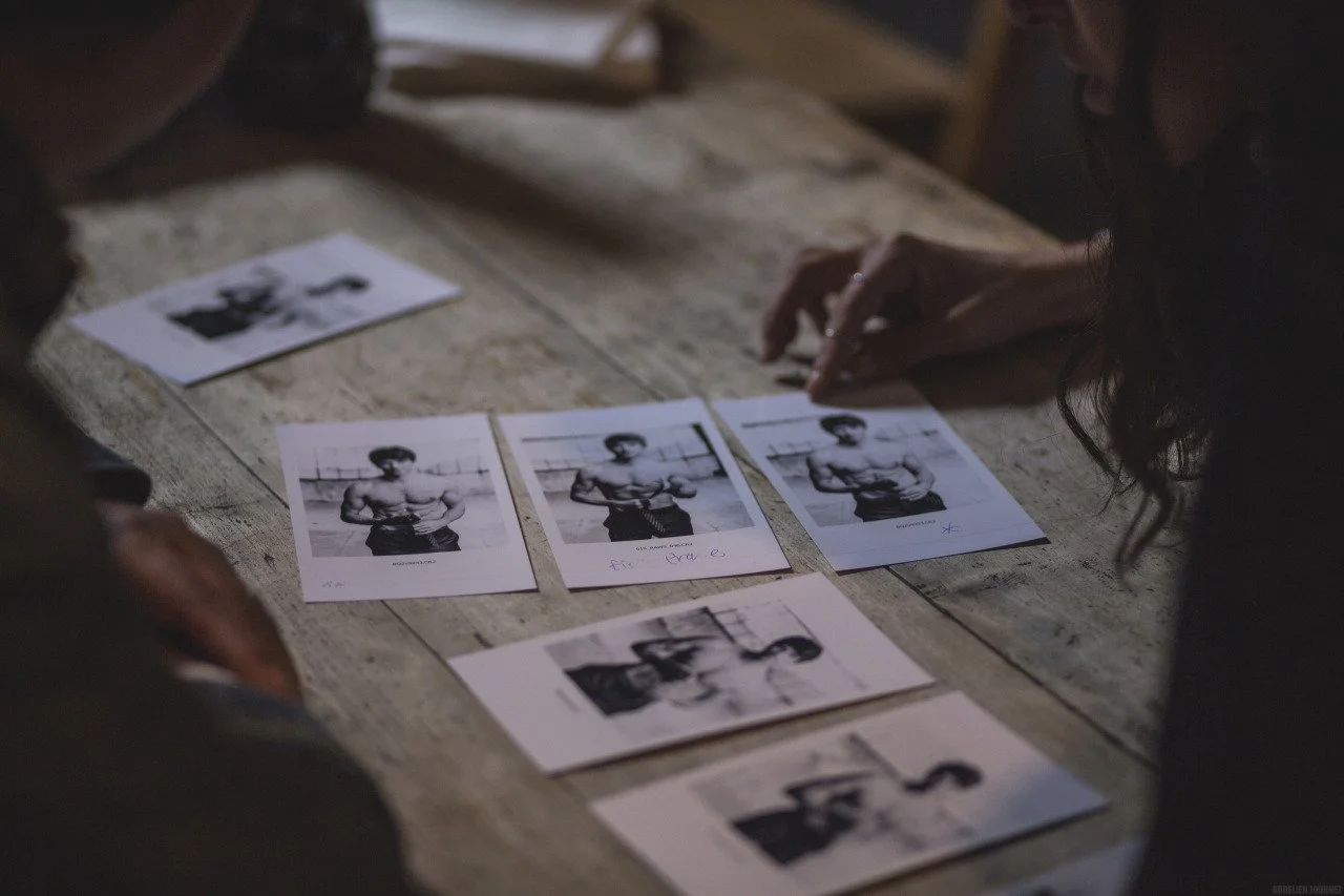 Deux personnes prennent des photos d'un homme musclé posant dans un gymnase, plusieurs photos sont disposées sur une table en bois.
