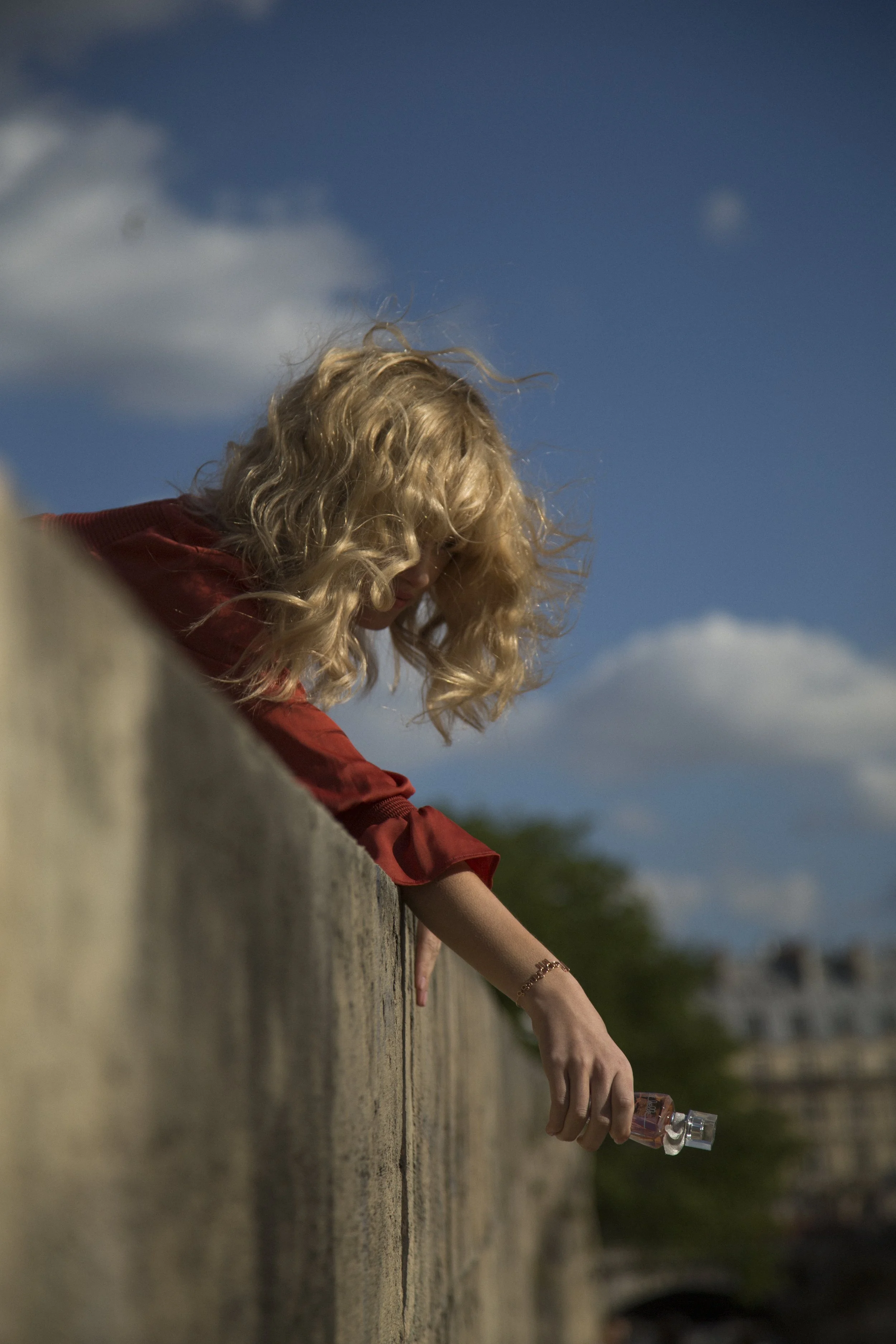 Fille blonde avec des cheveux bouclés qui regarde le sol, tenant une petite bouteille en verre, posée sur un vieux mur en pierre, avec un ciel bleu et des nuages en arrière-plan.