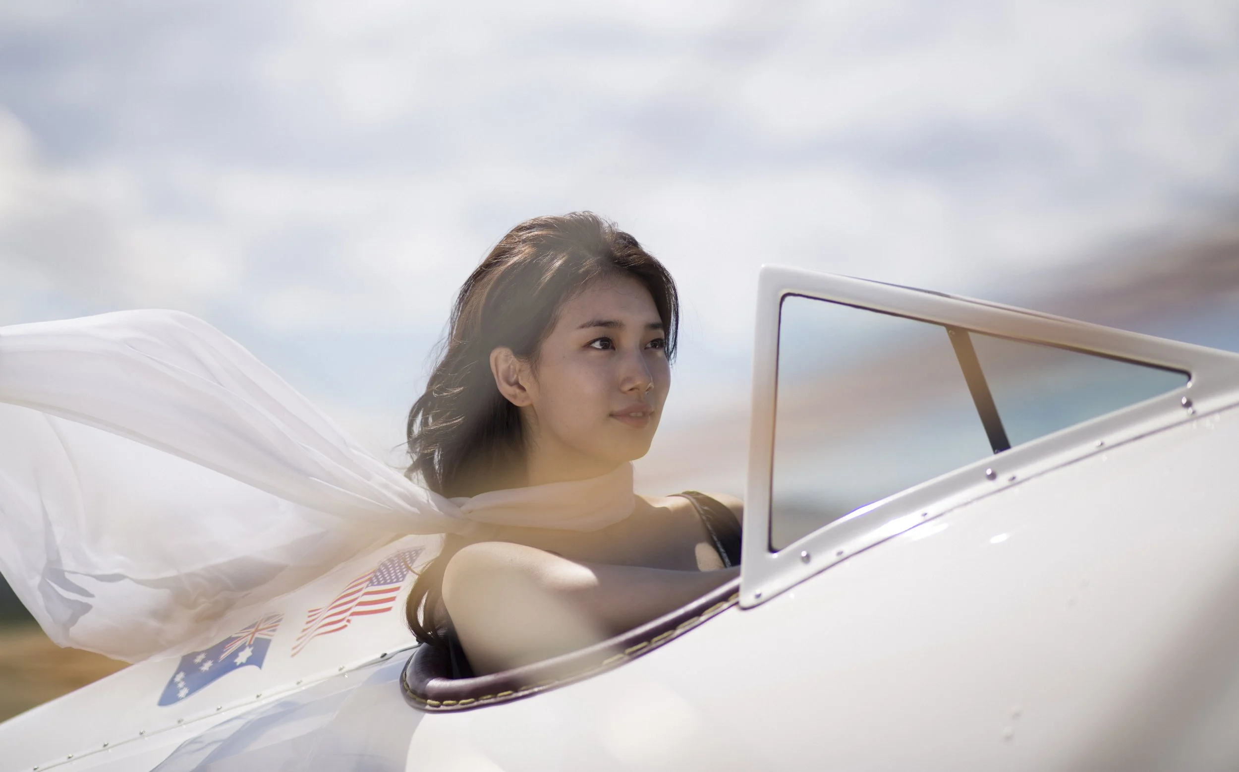 Une femme assise dans un cockpit à moitié ouvert d'un avion blanc, avec un drapeau américain et un drapeau australien visibles sur le côté, sous un ciel partiellement nuageux.