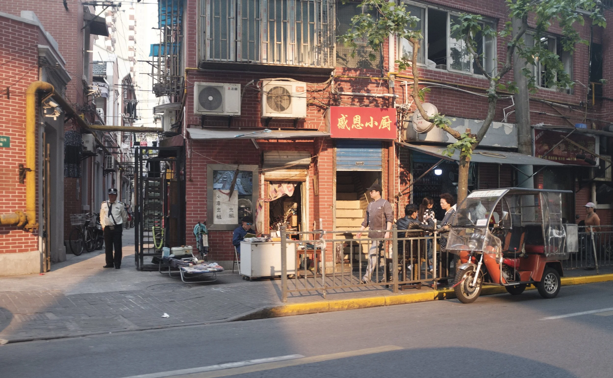 Commerce de rue avec des barrières en métal, un tuk-tuk rouge garé, des personnes qui discutent, une boutique avec un panneau en chinois, des appareils de climatisation sur un bâtiment en briques rouges, et une personne en uniforme de sécurité.