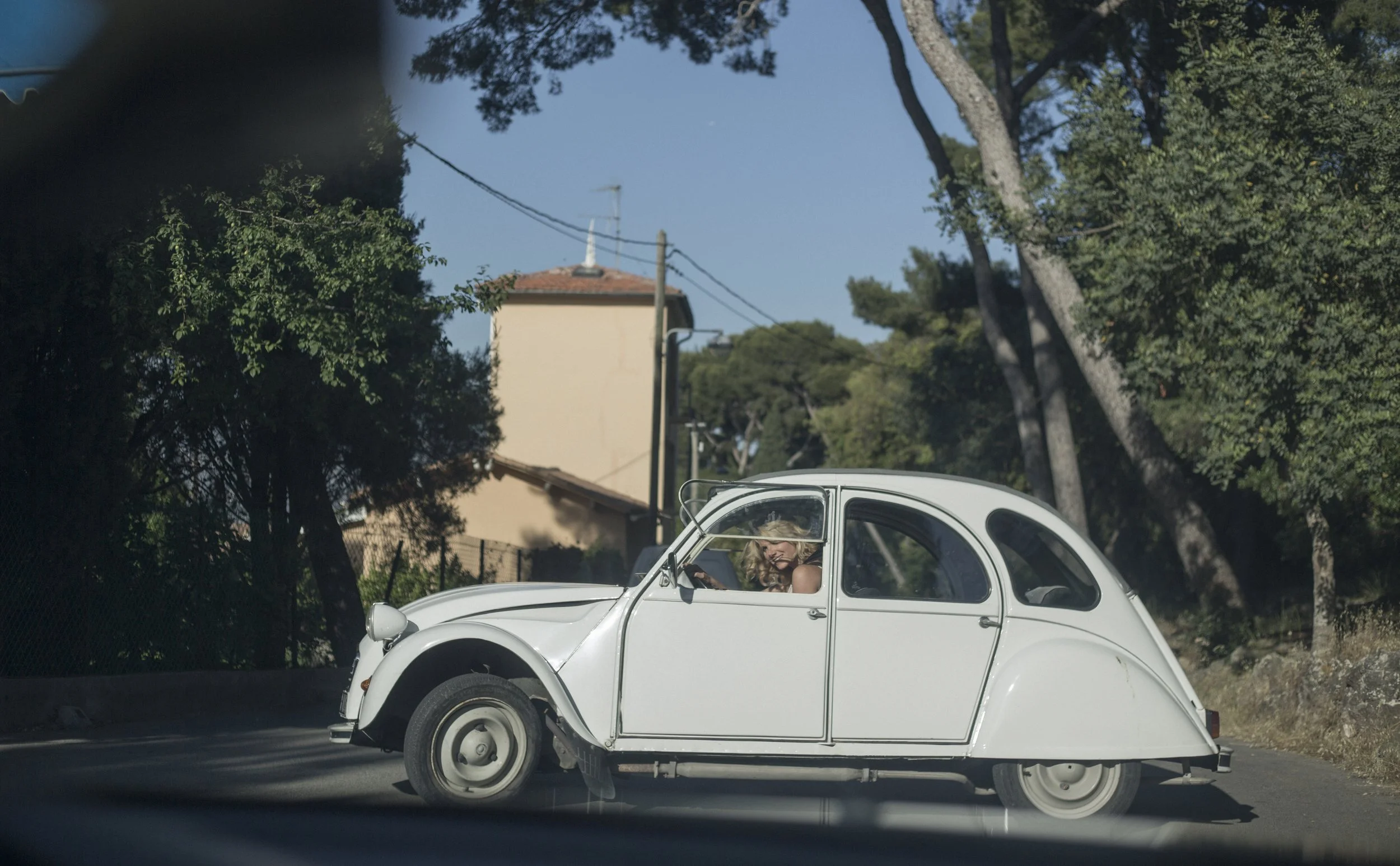Une voiture blanche de style Citroën 2CV sur une route, avec une femme à l'intérieur regardant par la fenêtre, entourée d'arbres et de maisons sous un ciel clair.
