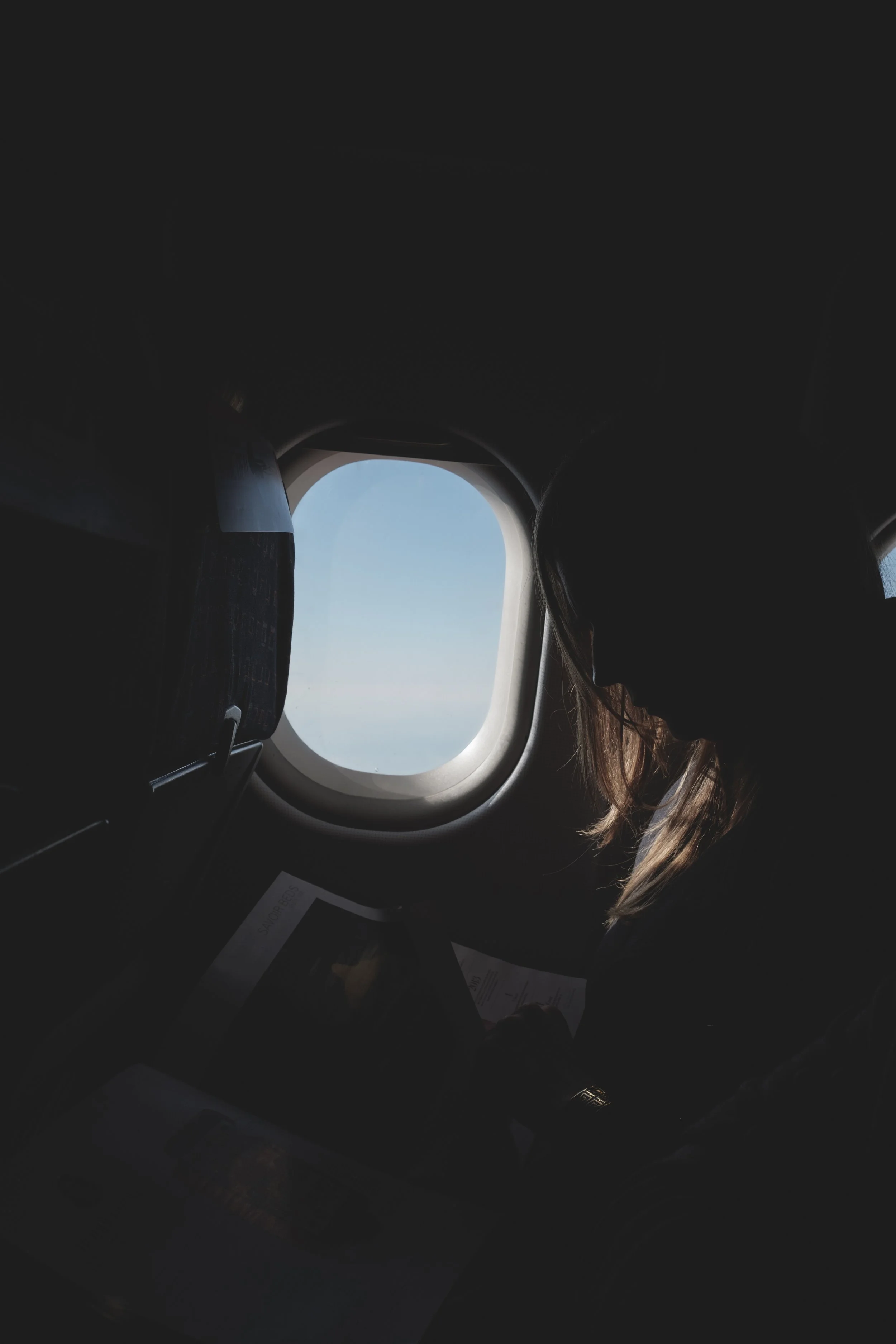 Une femme assise à une fenêtre d'avion, regardant un écran ou un document, avec le ciel bleu visible à travers la fenêtre.