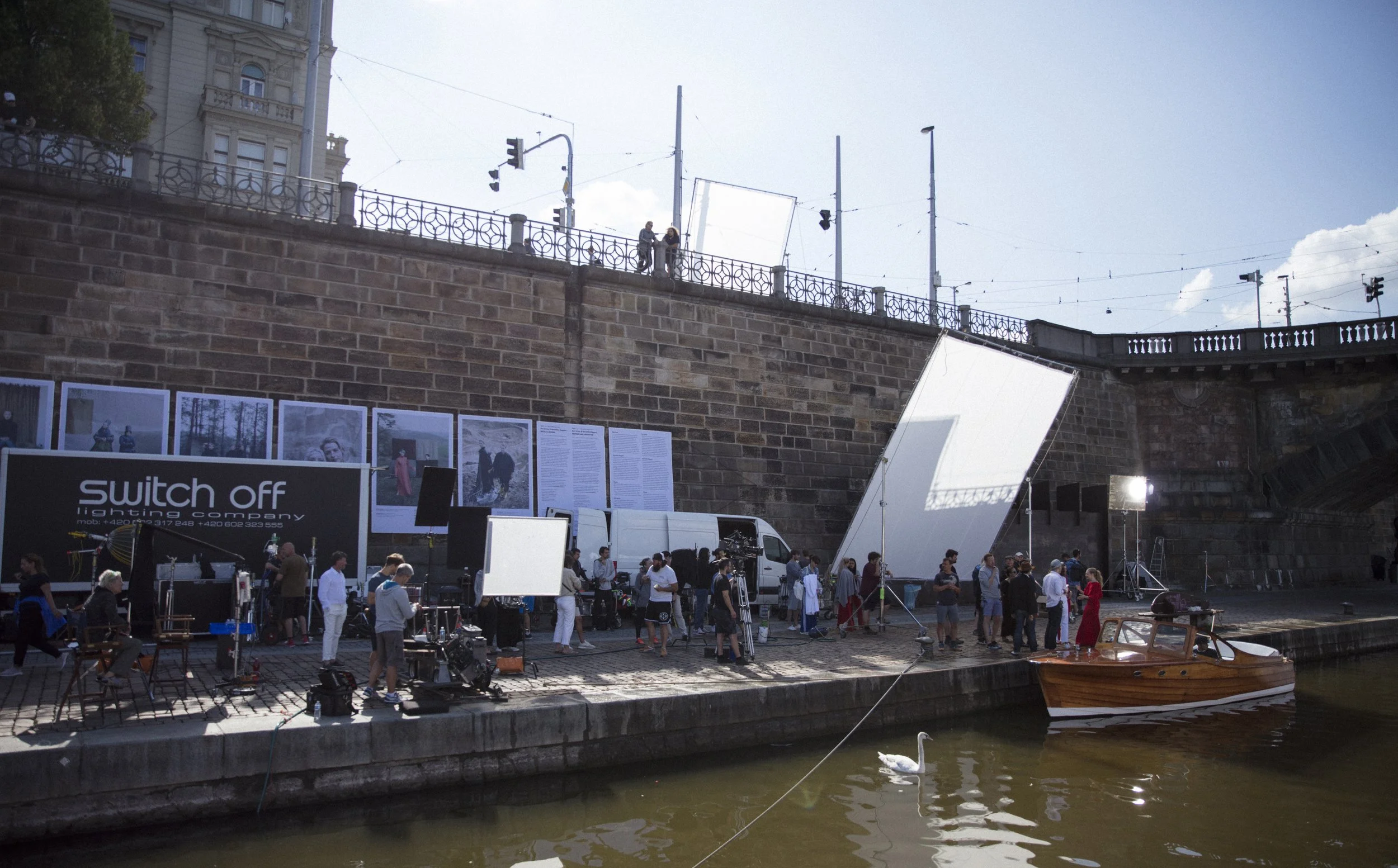 Une scène de tournage en extérieur au bord de l'eau avec des personnes, des équipements de film, un petit bateau en bois, et un pont en pierre en arrière-plan.