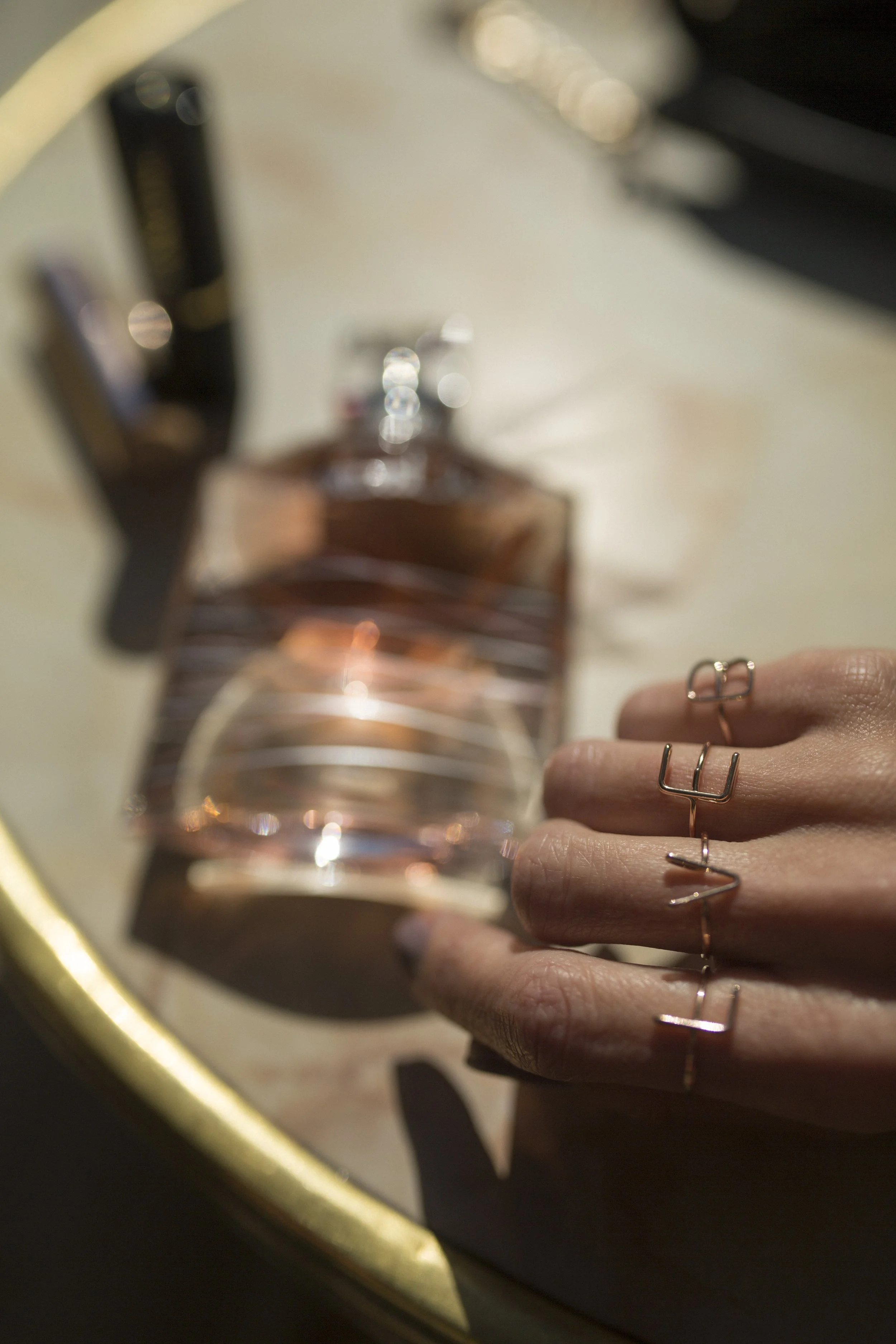 Une main avec des bagues repose sur une surface en face d'un flacon de parfum, avec une bougie allumée et d'autres objets flous en arrière-plan.
