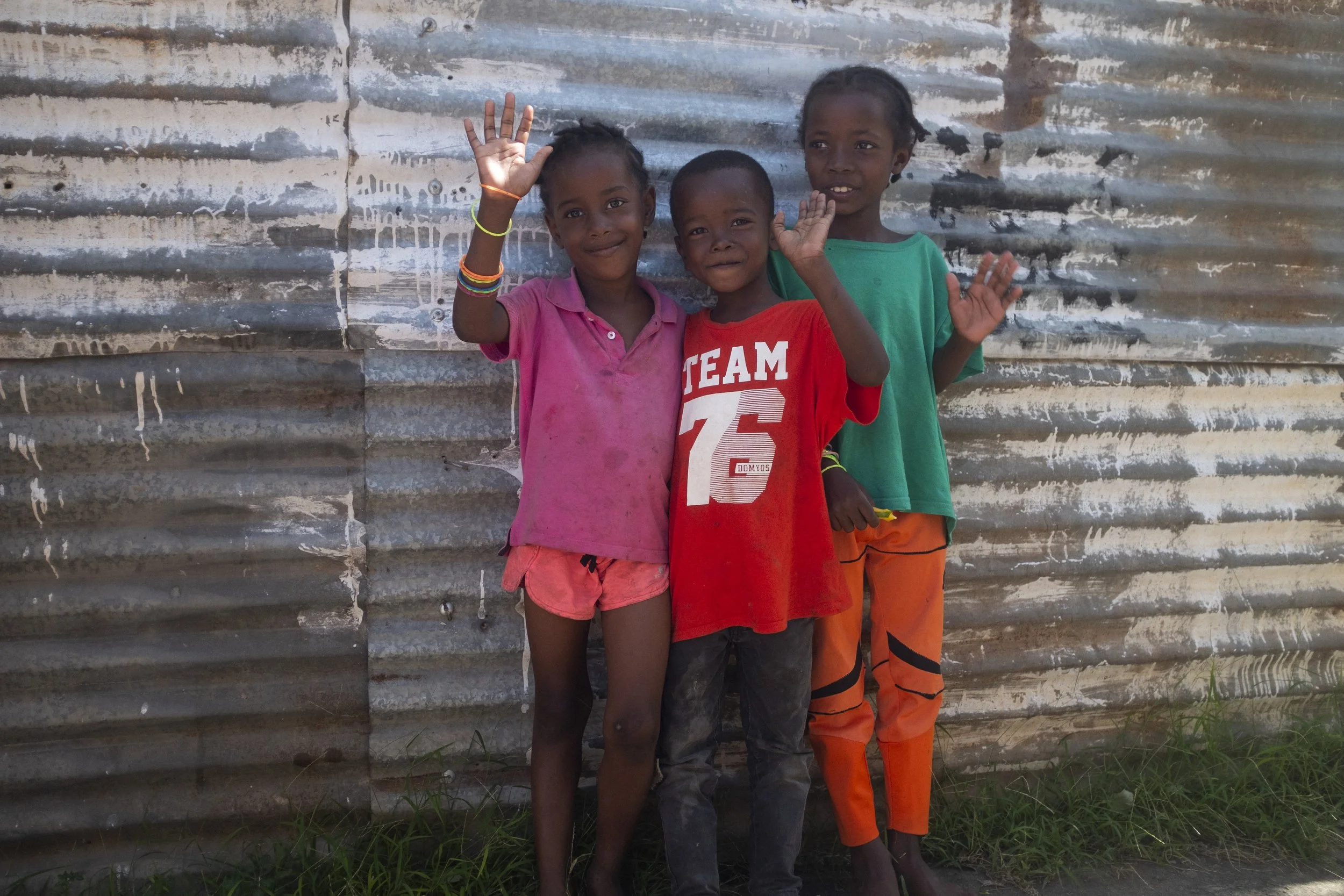 Trois enfants souriants debout devant un mur en tôle ondulée, saluant la caméra, dans un environnement extérieur.