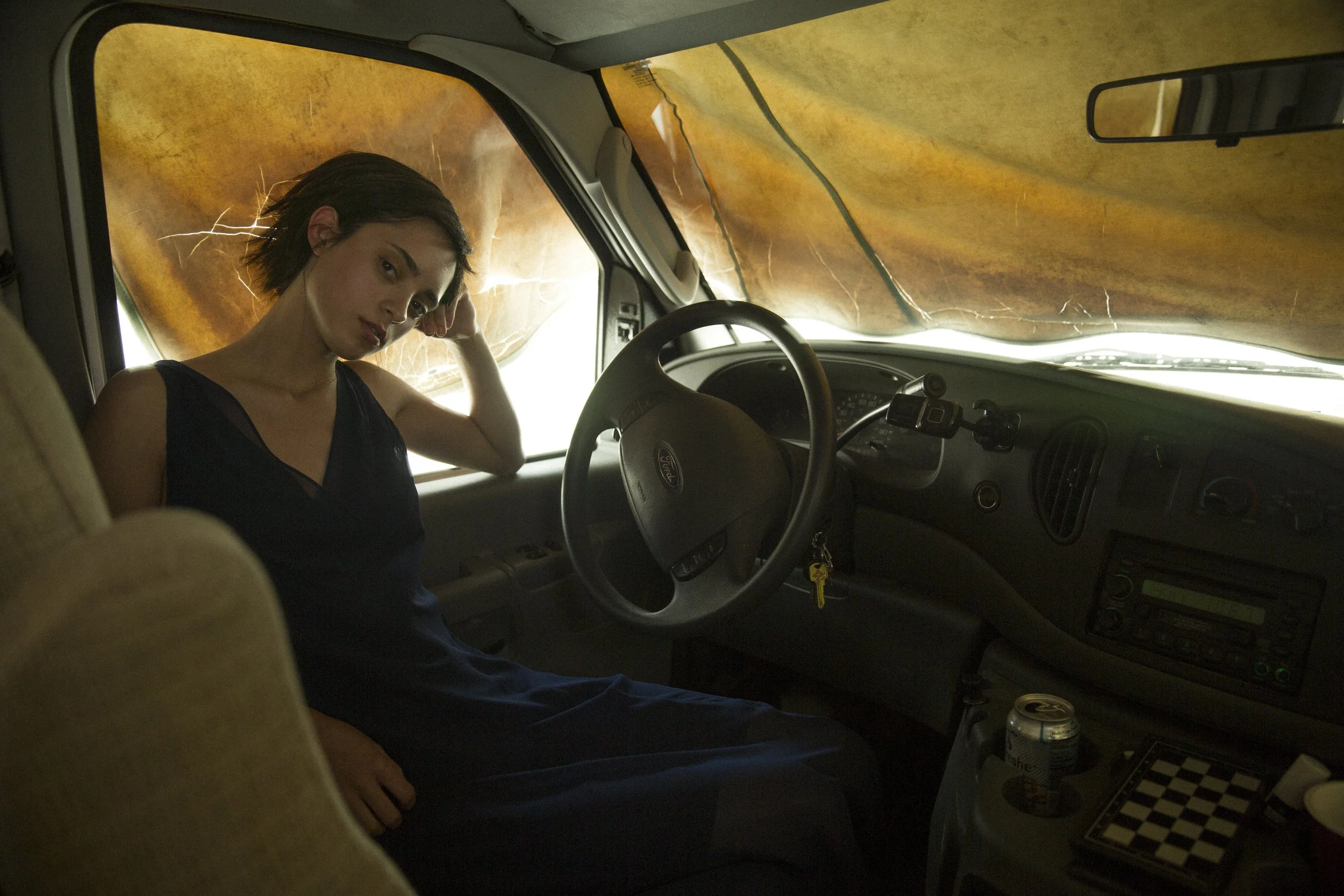 Une femme assise sur le siège conducteur d'un véhicule, avec un volet orange suspendu à la fenêtre derrière elle, et divers objets sur le tableau de bord, notamment une canette, un échiquier et un porte-clés.