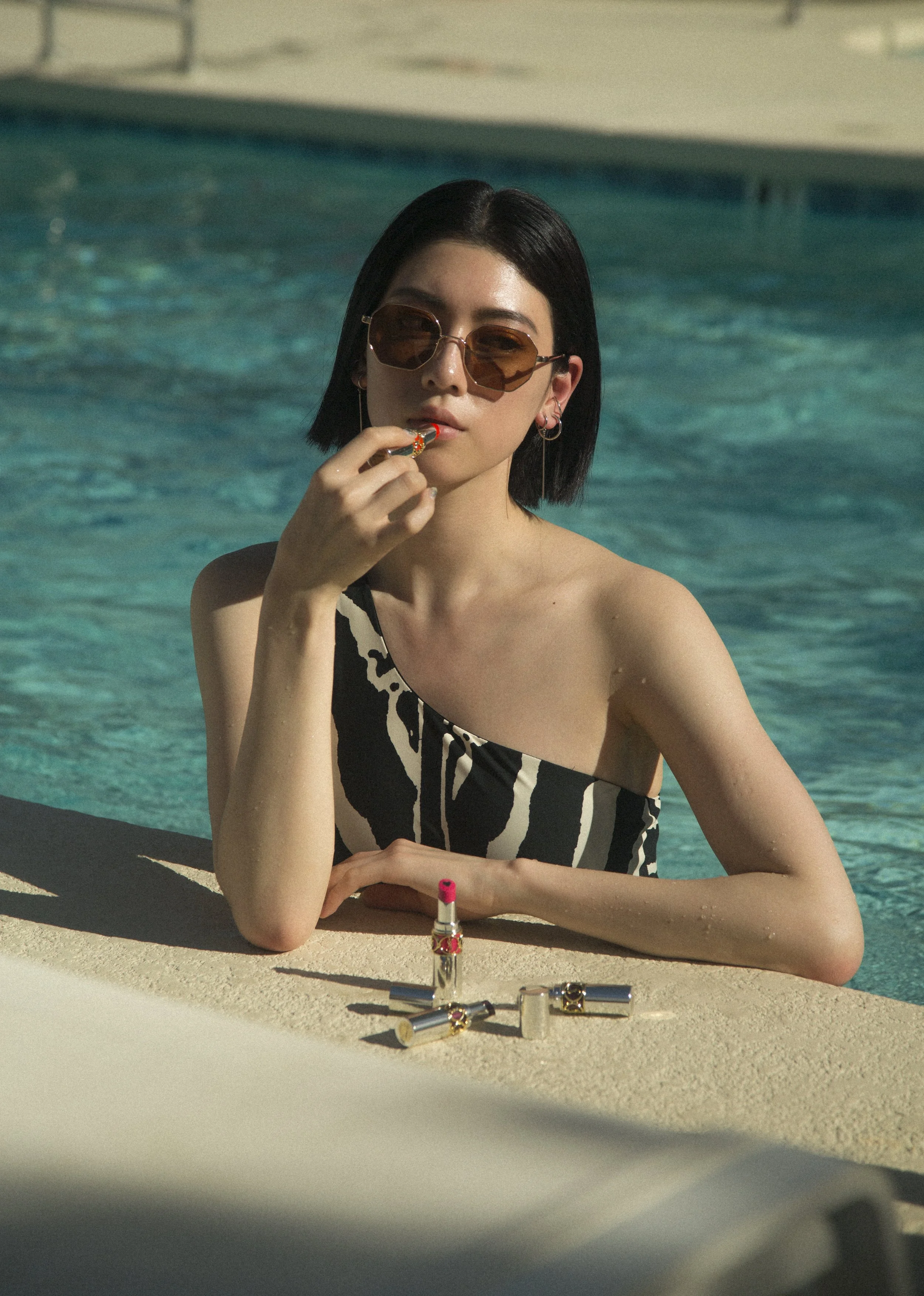 Une femme brune portant des lunettes de soleil, une robe rayée noire et blanche, est assise au bord d'une piscine, en train de mettre du rouge à lèvres.