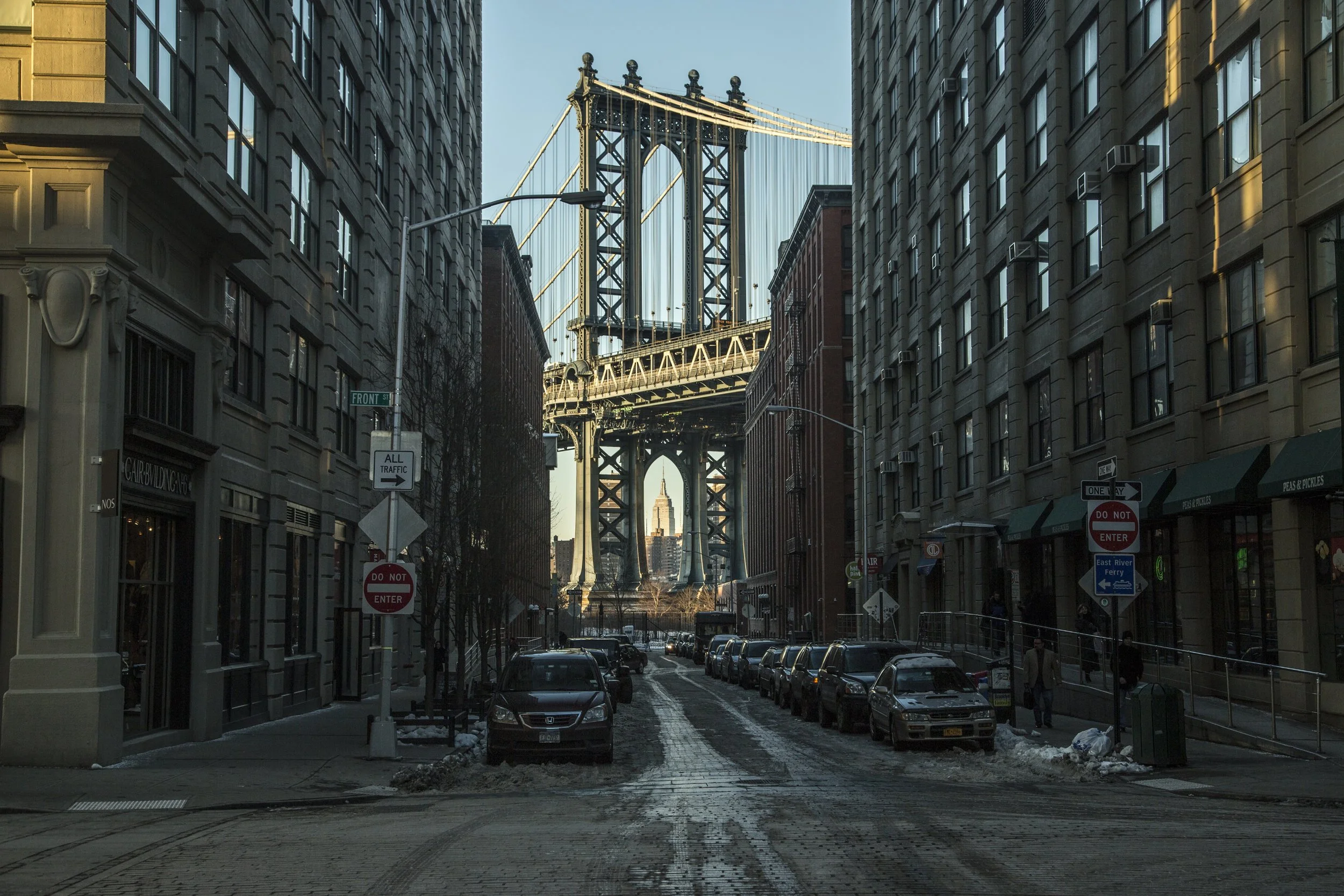 Vue urbaine de la rue avec des voitures garées, entourée de bâtiments avec le pont de Brooklyn en arrière-plan et l'empire State Building visible au loin.