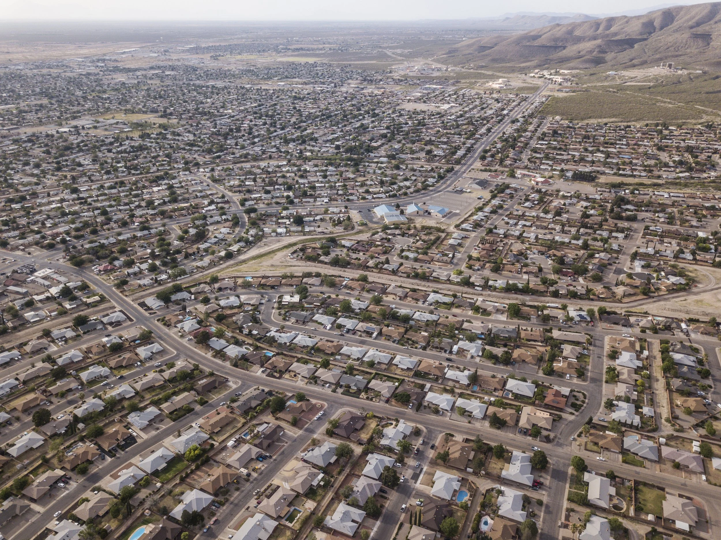 Vue aérienne d'un quartier résidentiel avec de nombreuses maisons, routes et quelques espaces verts, entouré de montagnes en arrière-plan.