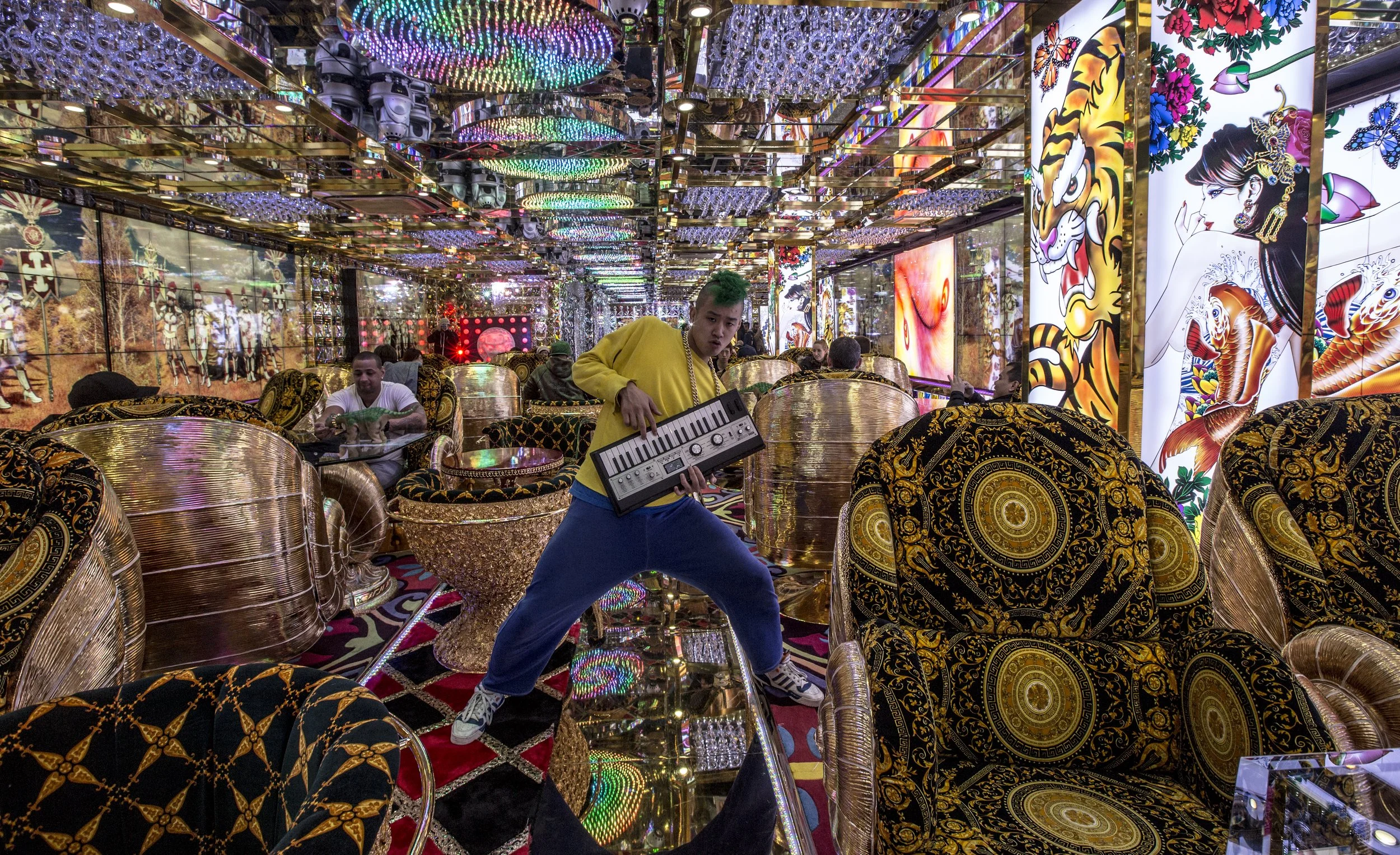 Jeune homme avec une couleur de cheveux verte, portant un sweat jaune et un pantalon bleu, jouant un clavier électronique dans un décor luxueux et coloré avec des sièges et des murs ornés de motifs or et de grandes images de tigres, de femmes et de f