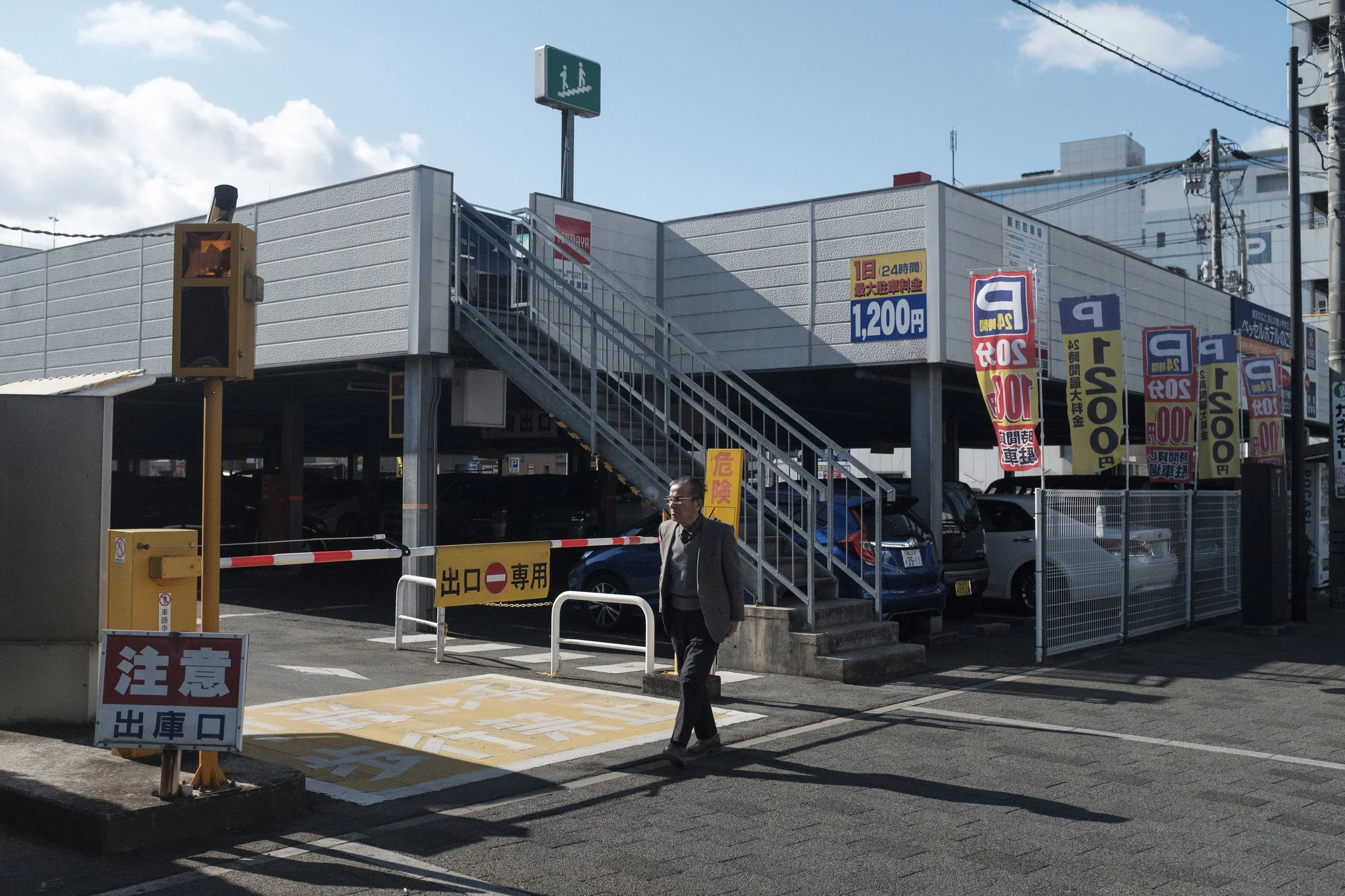 Parking extérieur avec un homme marchant près d'une barrière rouge et blanche, un panneau jaune avec des caractères japonais, des voitures stationnées sous un niveau supérieur accessible par un escalier en métal, plusieurs banderoles jaunes avec des 