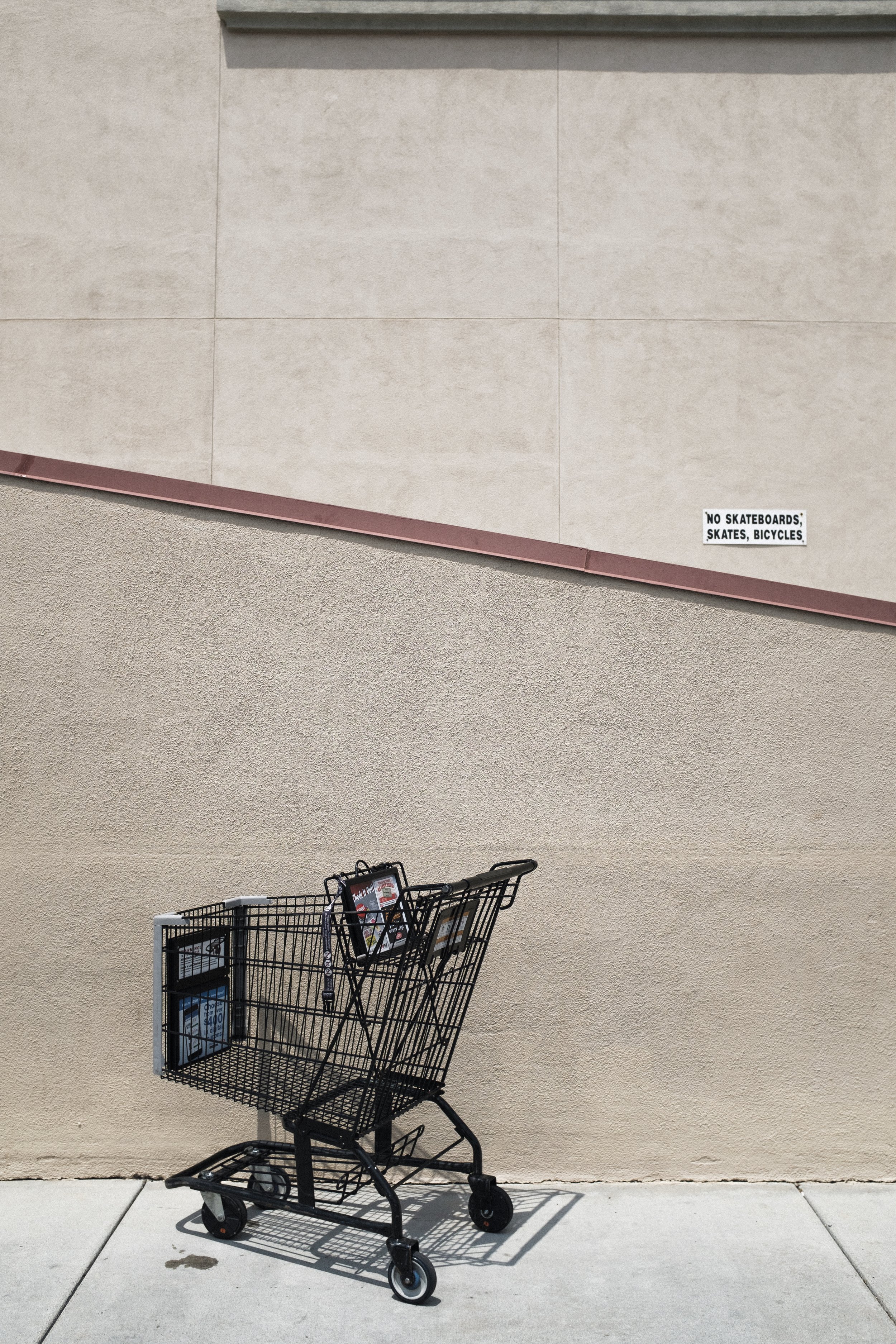 Panier de supermarché vide placé sur un trottoir devant un mur beige avec un panneau blanc indiquant 'No skateboards, skates, bicycles'.