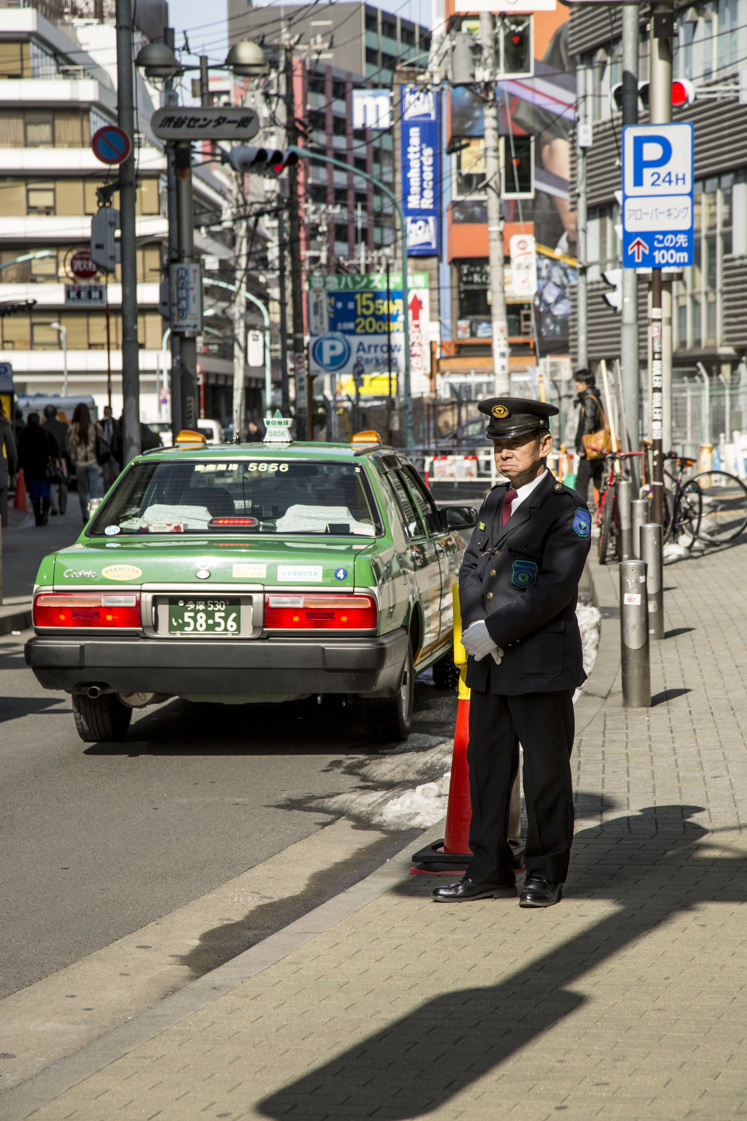 Une scène urbaine au Japon avec un agent de police en uniforme et un taxi vert stationnés sur le côté de la rue. Il y a des panneaux de signalisation, des bâtiments et des passants en arrière-plan.