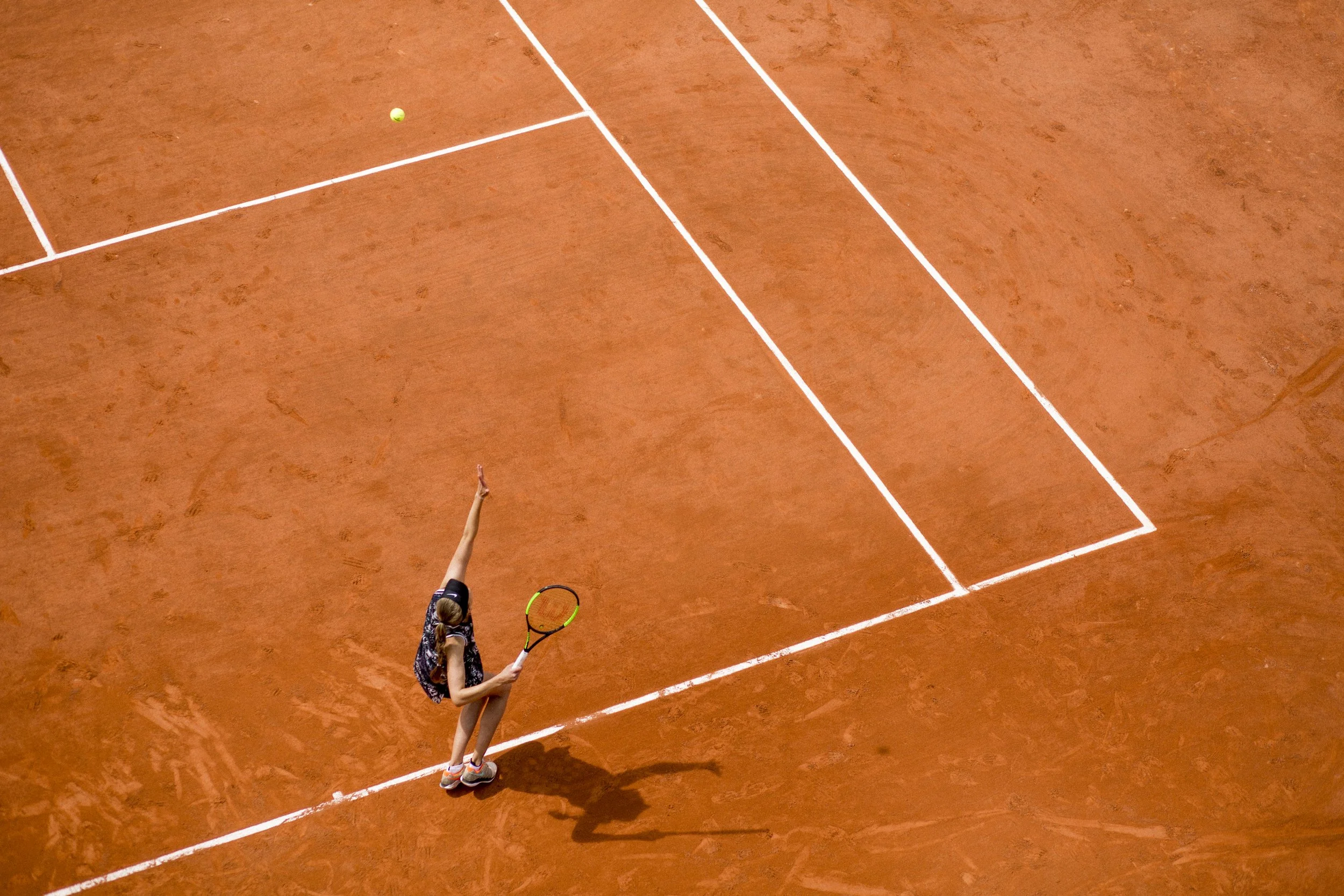 Une joueuse de tennis féminine sur un court en terre battue, portant une tenue noire et une visière, préparant un service.