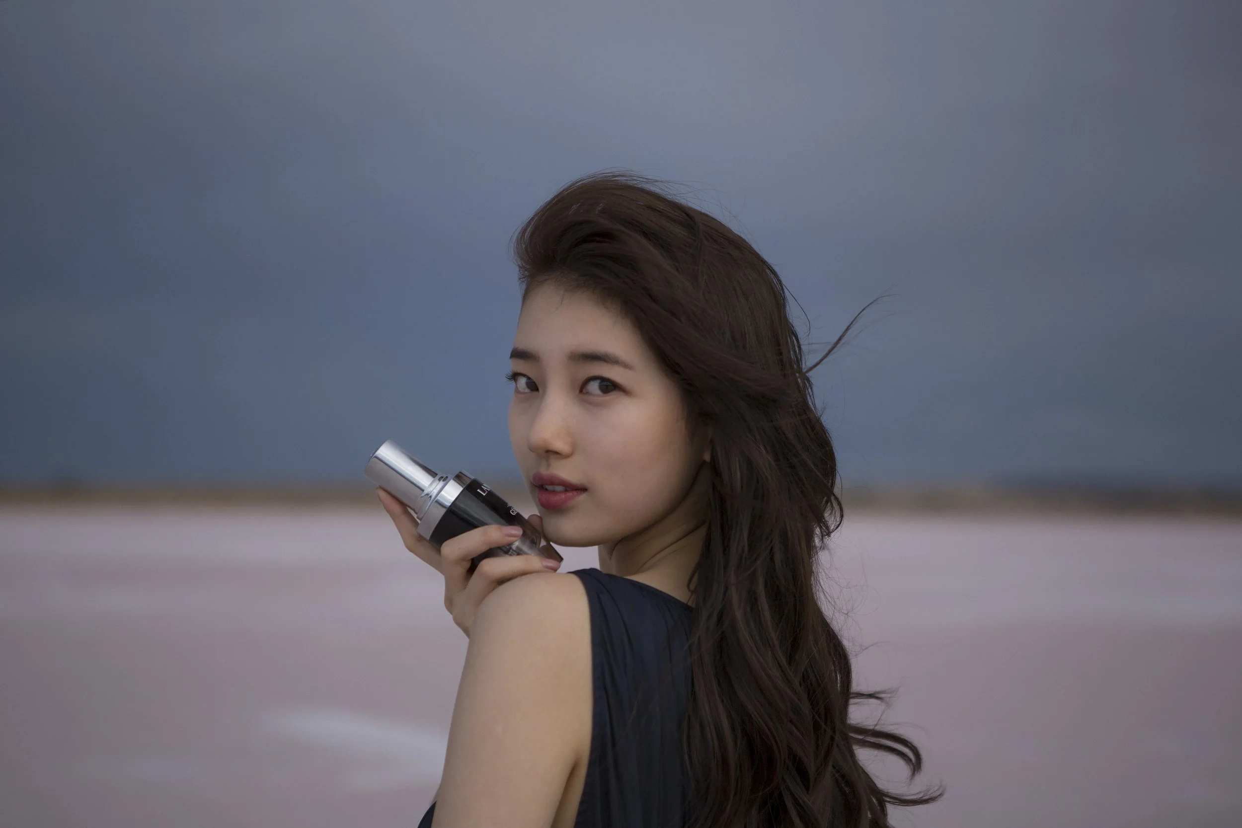 Une jeune femme aux longs cheveux bruns regarde par-dessus son épaule, tenant un appareil photo devant un paysage côtier avec un ciel nuageux.