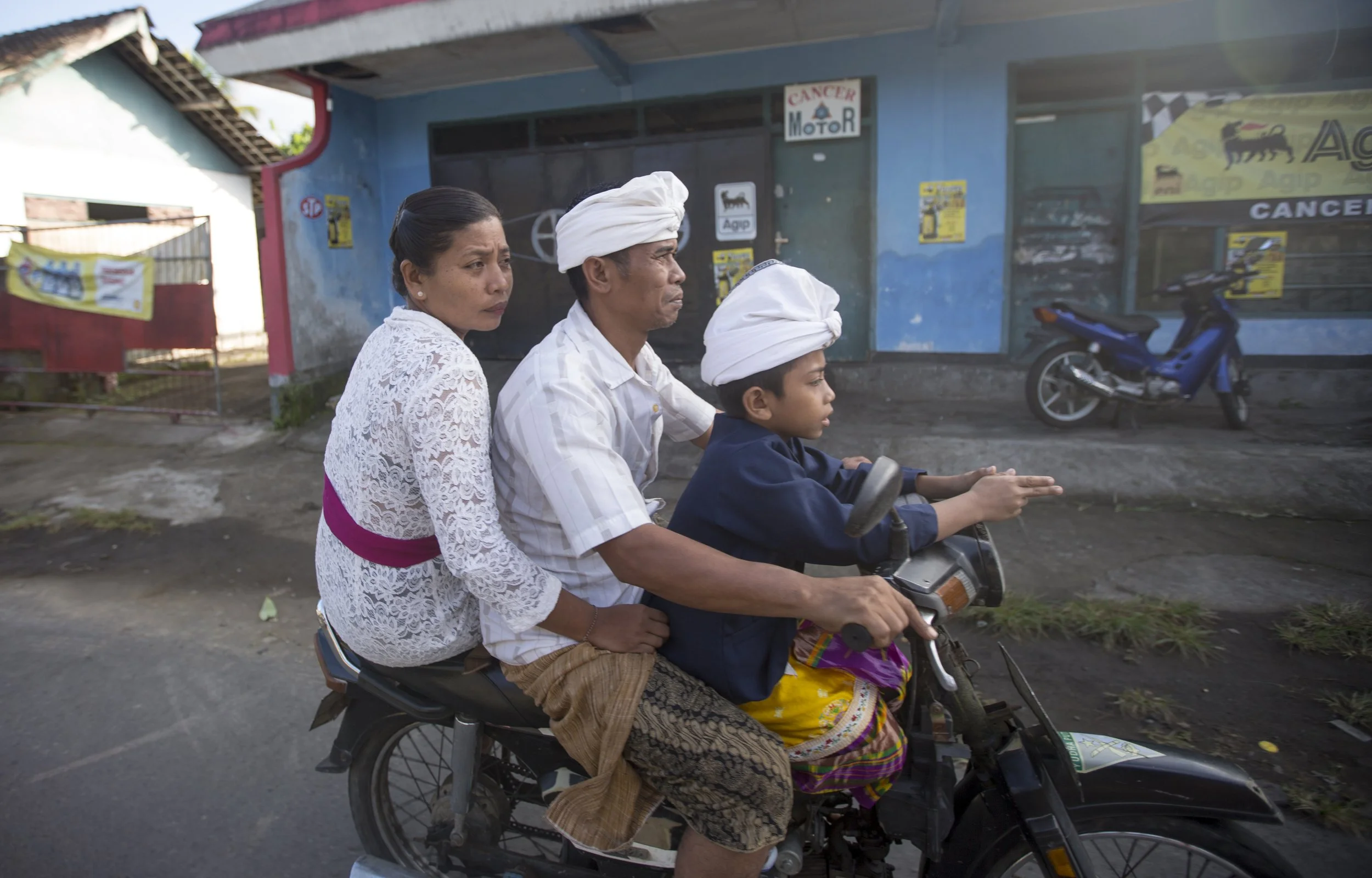 Une famille voyageant en moto : un homme, une femme et un garçon, tous portant des turbans blancs, en route dans une rue urbaine.