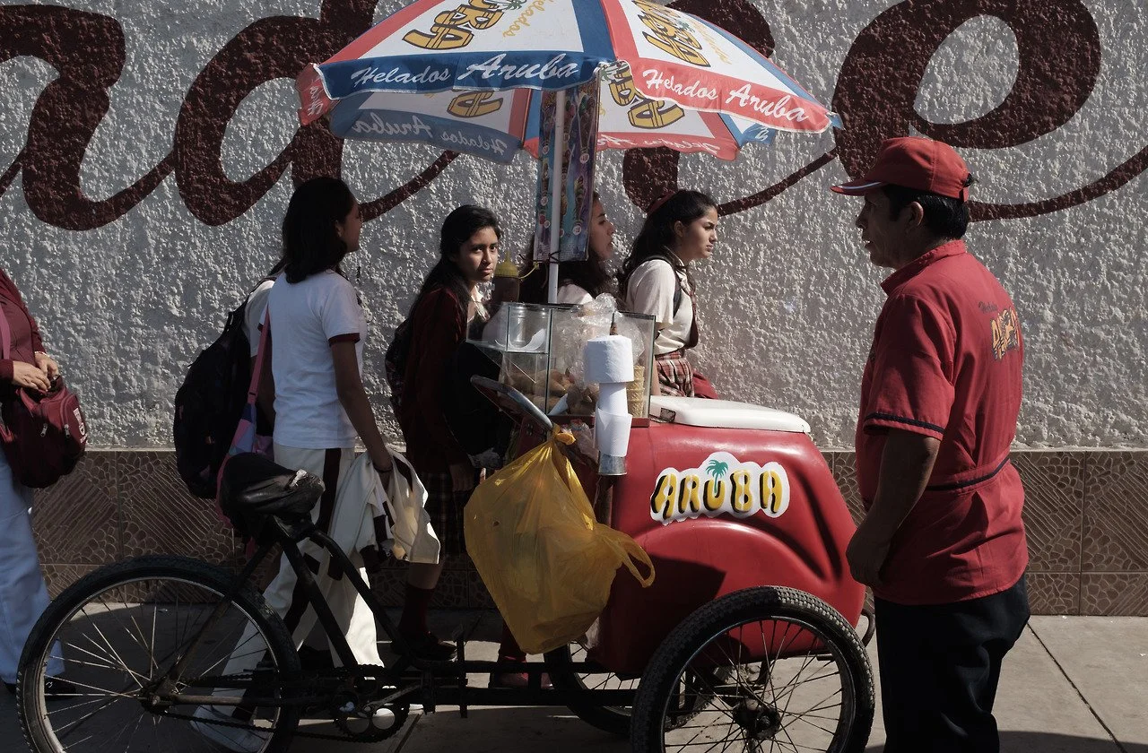 Vendeur de glaces sur un triporteur en interaction avec des jeunes filles, avec un parasol coloré et un mur en arrière-plan