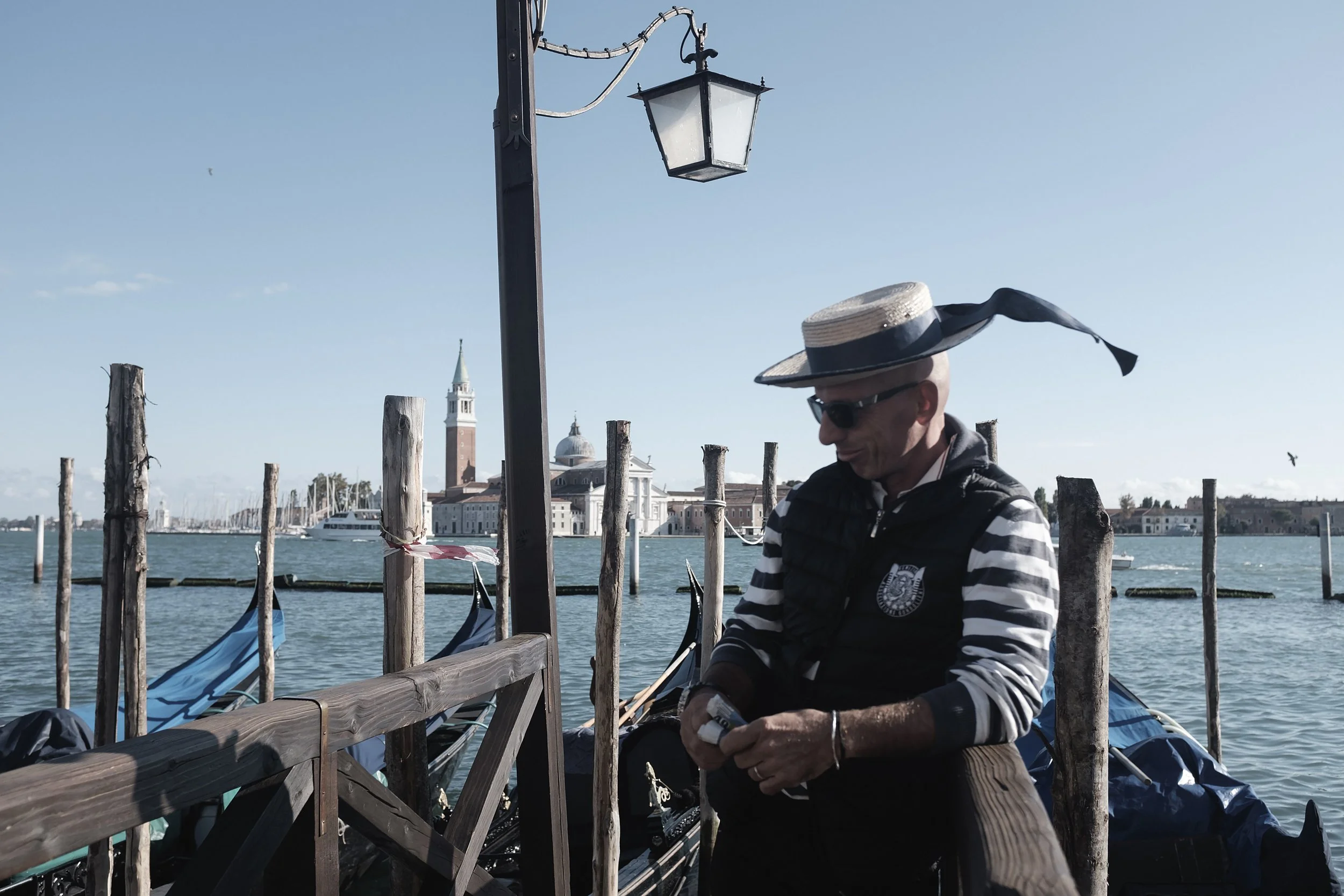 Un homme portant un chapeau large, des lunettes de soleil, une veste noire et une chemise à rayures assis sur une jetée en bois à Venise, avec un canal, des gondoles, un clocher et des bâtiments historiques en arrière-plan.