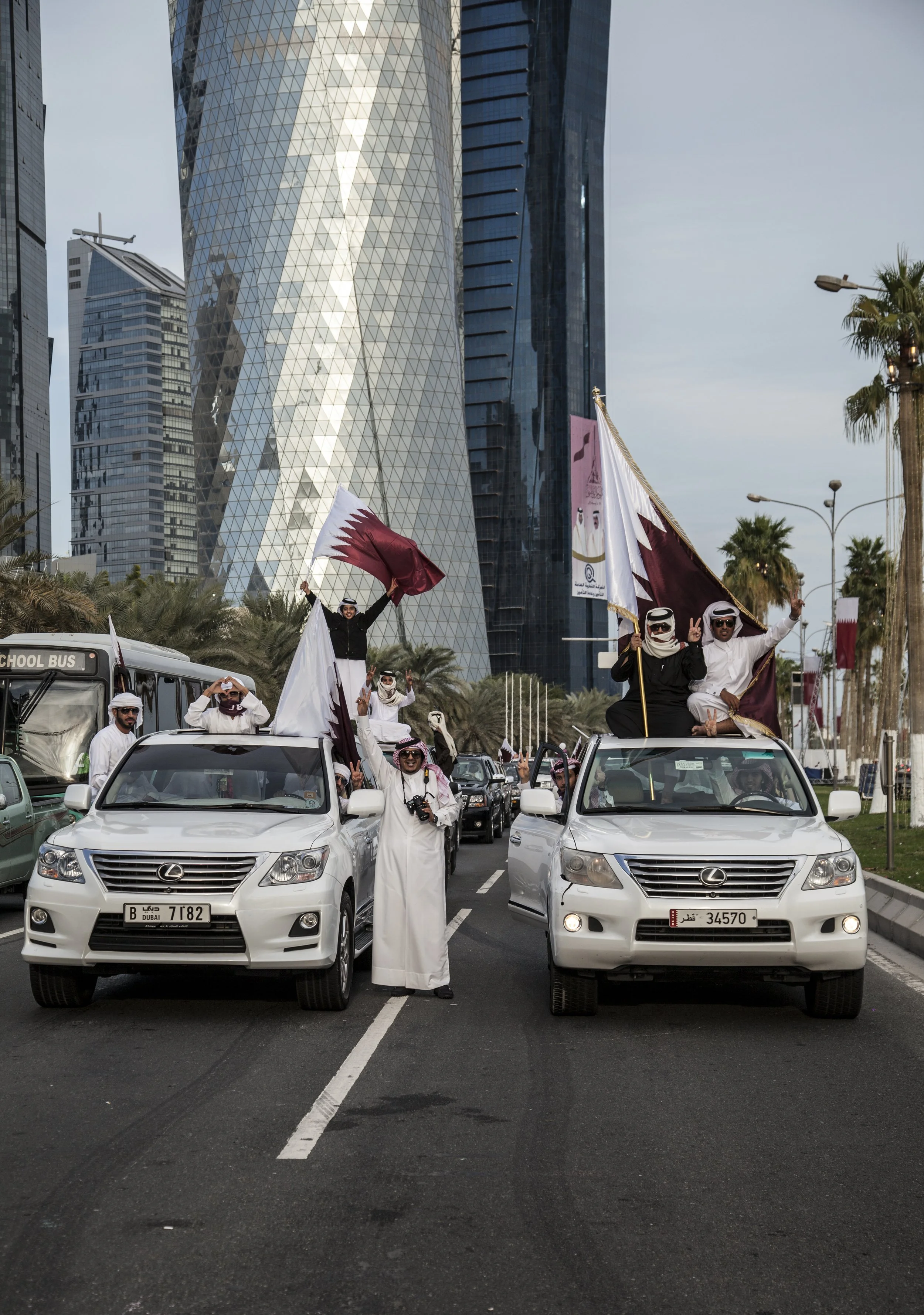 Groupe de personnes en vêtements traditionnels arabes célébrant, tenant des drapeaux du Qatar, lors d'une parade en voiture avec des gratte-ciel modernes en arrière-plan à Doha, Qatar.