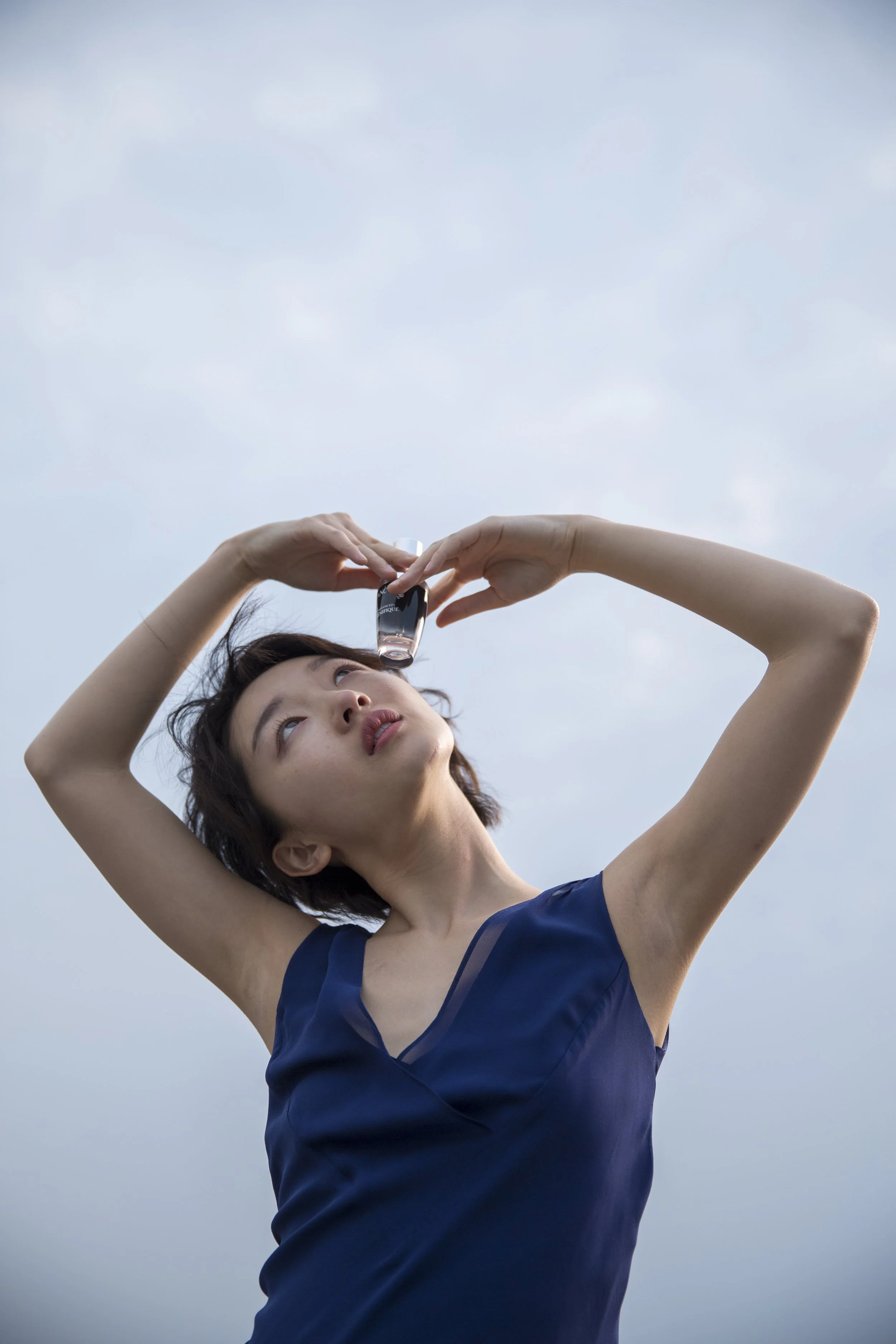 Une jeune femme tenant un flacon de parfum au-dessus de sa tête, regardant vers le haut, avec un ciel gris en arrière-plan.