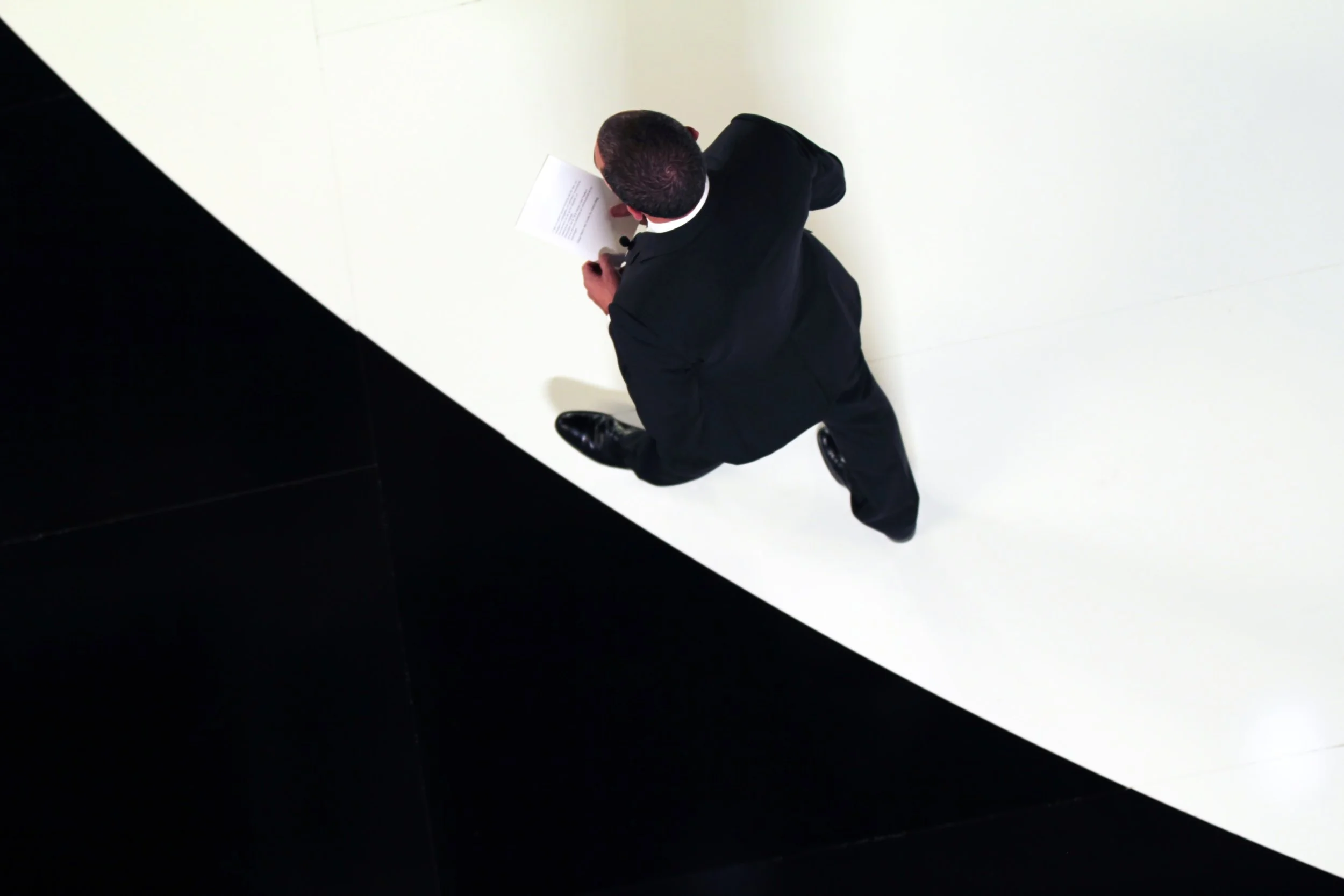 Vue aérienne d'un homme en costume noir assis sur un sol blanc, lisant un document, avec un fond noir en diagonale.