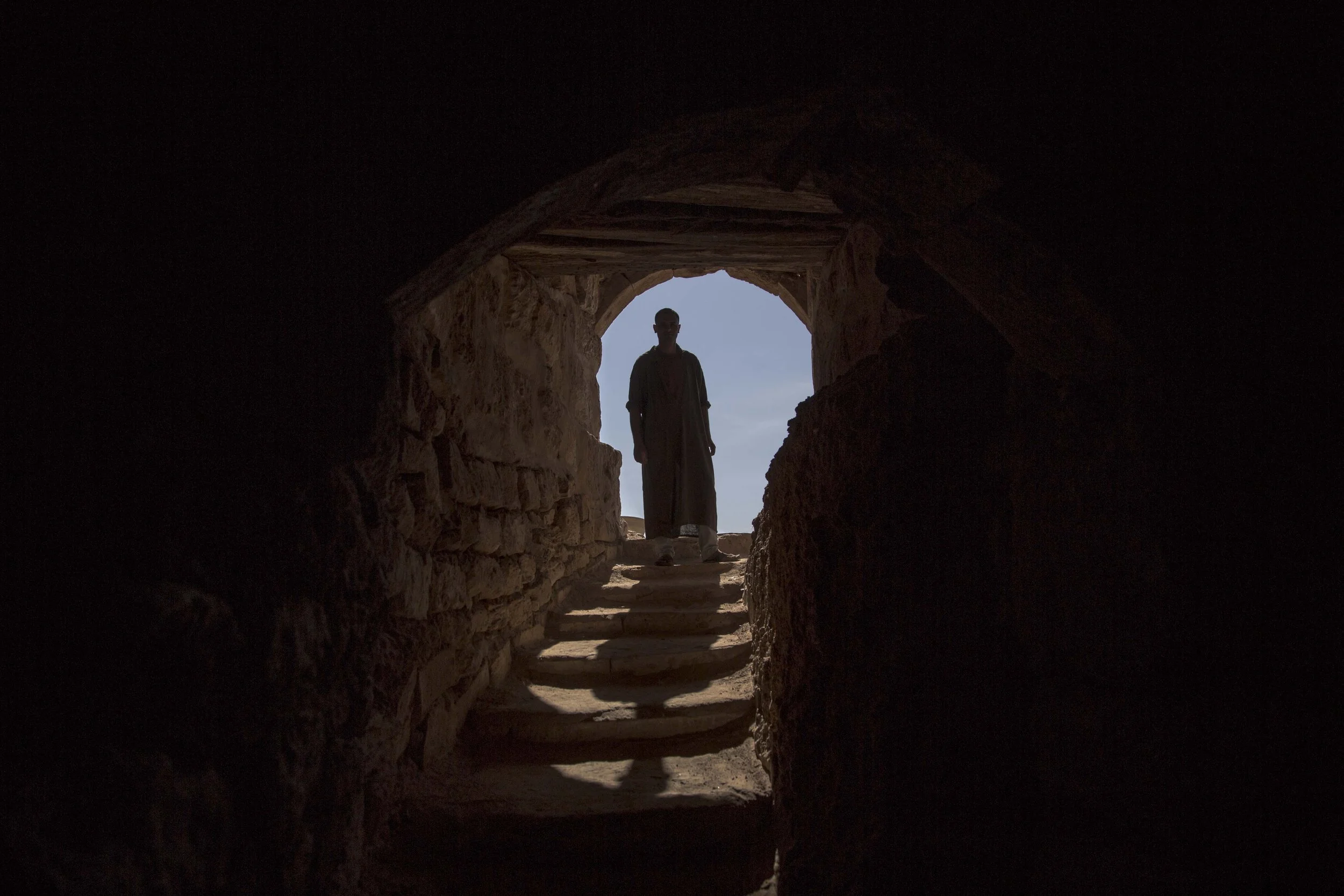Silhouette d'une personne debout à l'entrée d'une structure en pierre, vue depuis l'intérieur sombre vers un ciel bleu clair.