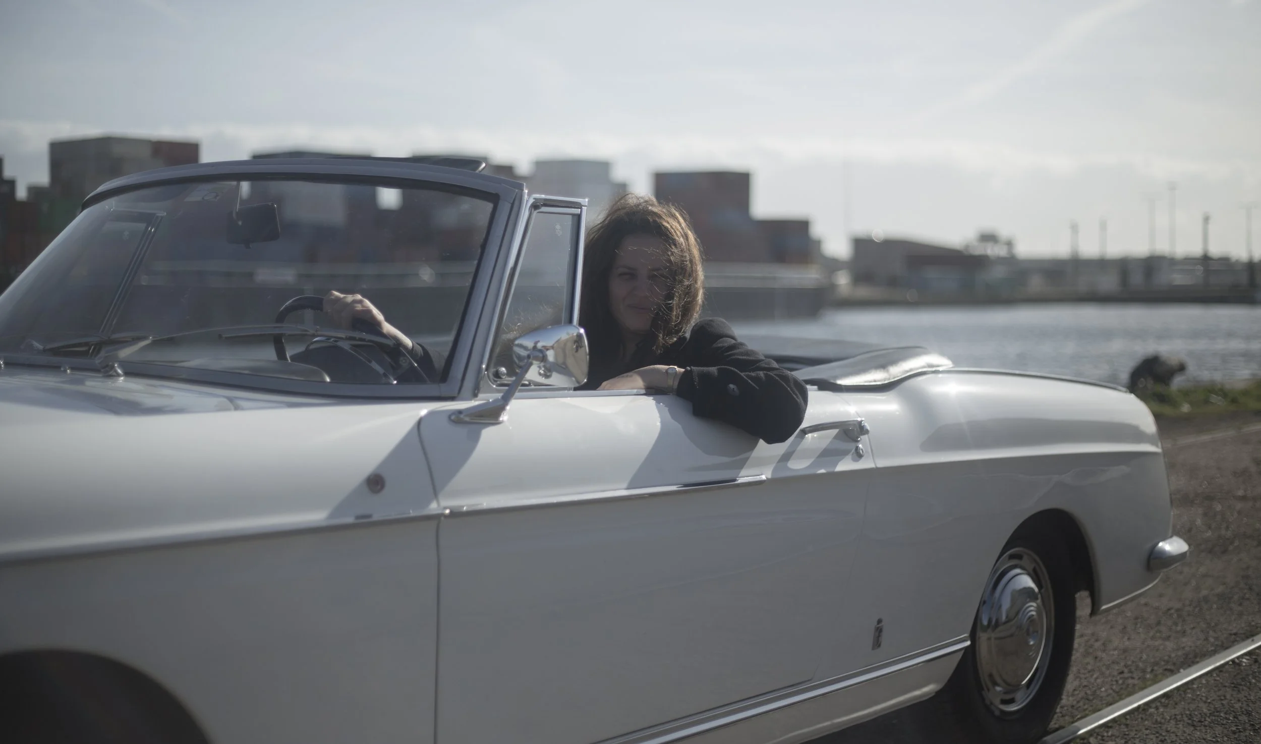 Une femme avec des cheveux bruns courts, assise dans une voiture décapotable blanche, près d'une rivière ou d'un plan d'eau, avec un paysage urbain en arrière-plan.