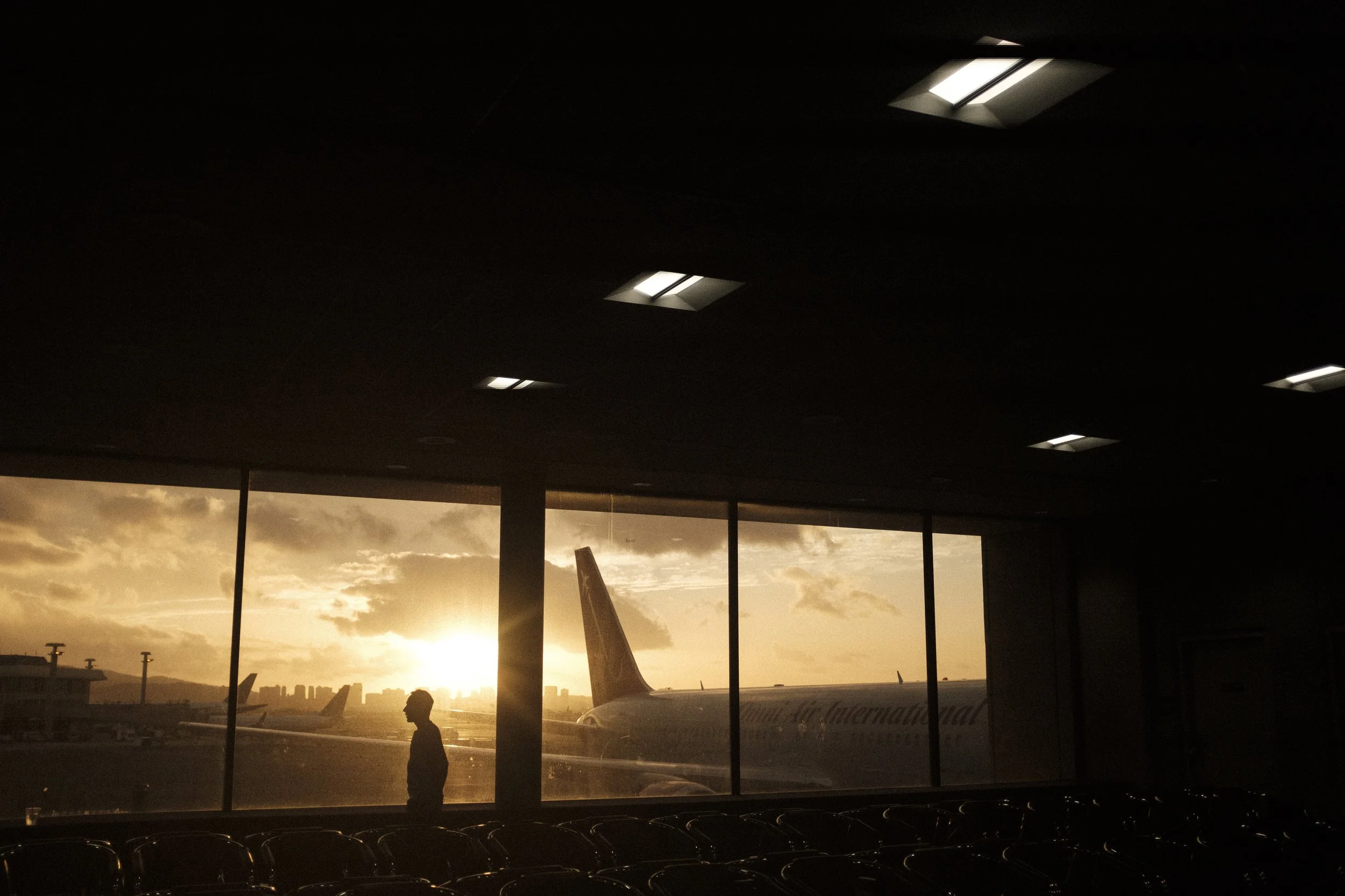 Silhouette d'une personne dans un aéroport, avec un avion visible par la fenêtre, lors d'un coucher de soleil.