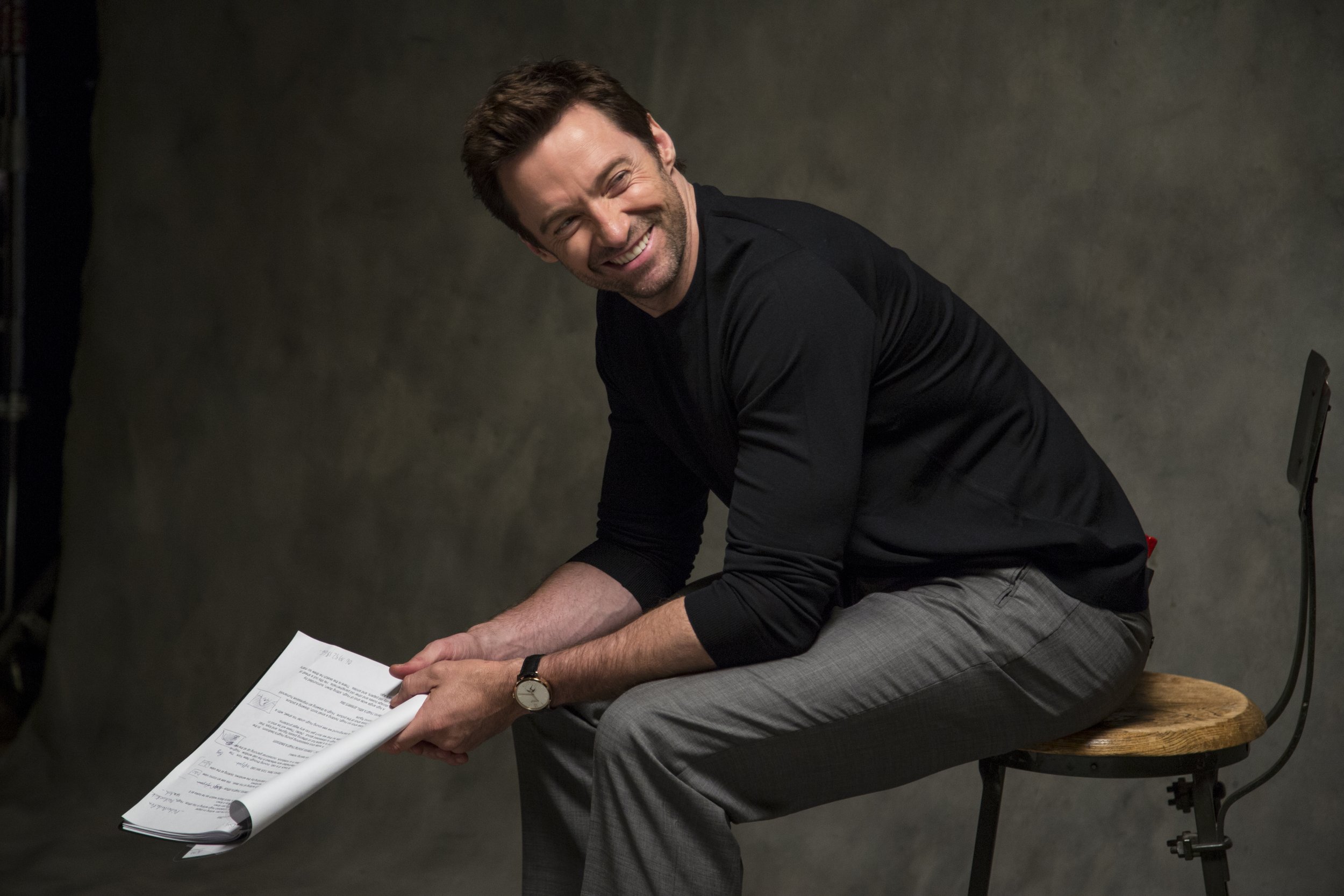 Un homme souriant, assis sur une chaise en bois, en tenue professionnelle avec un pantalon à carreaux gris et un pull noir, tenant des papiers dans ses mains, avec un fond sombre et neutre.