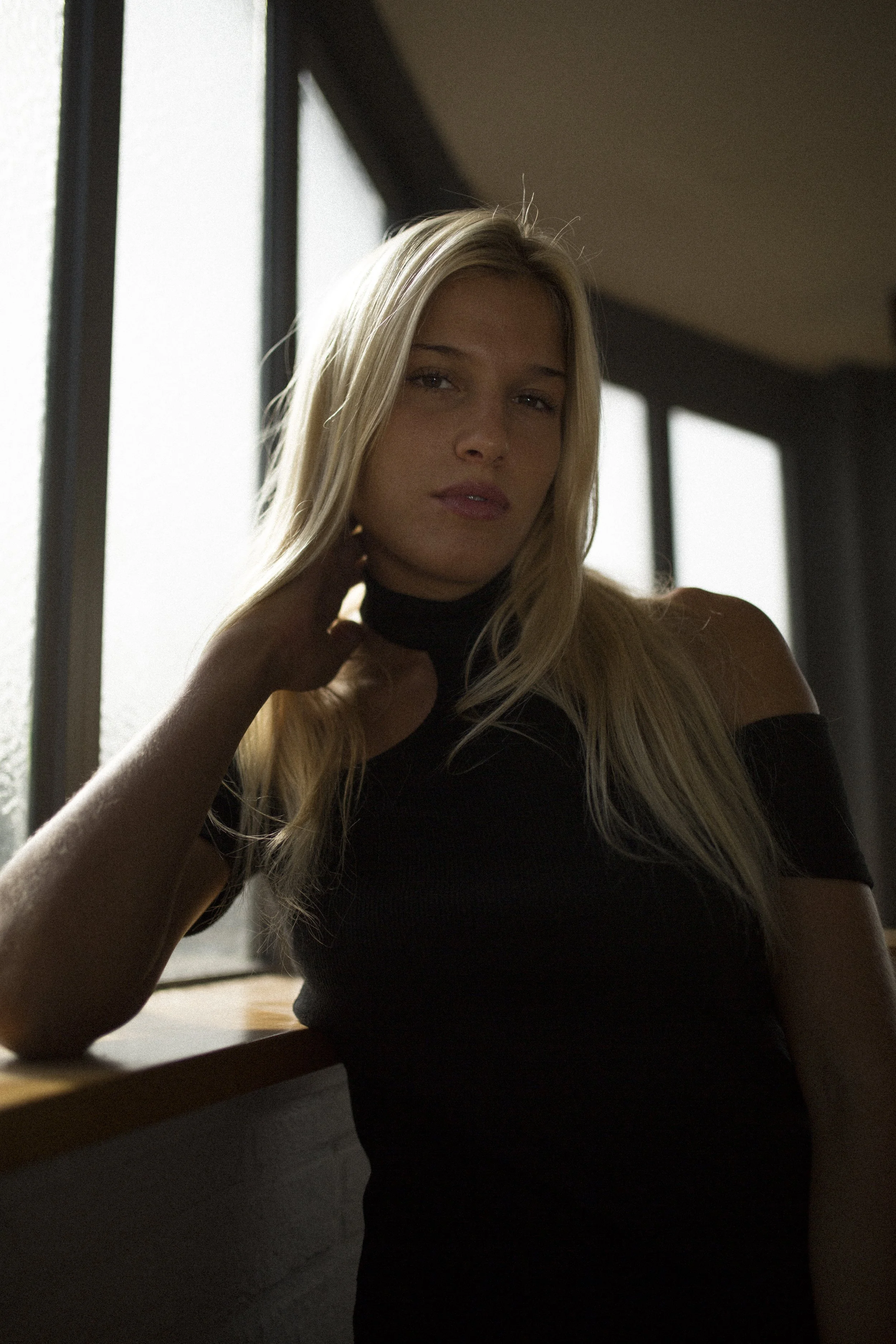 Femme aux cheveux longs, blonde, portant un haut noir avec épaule dénudée, posant près d'une fenêtre avec lumière naturelle.
