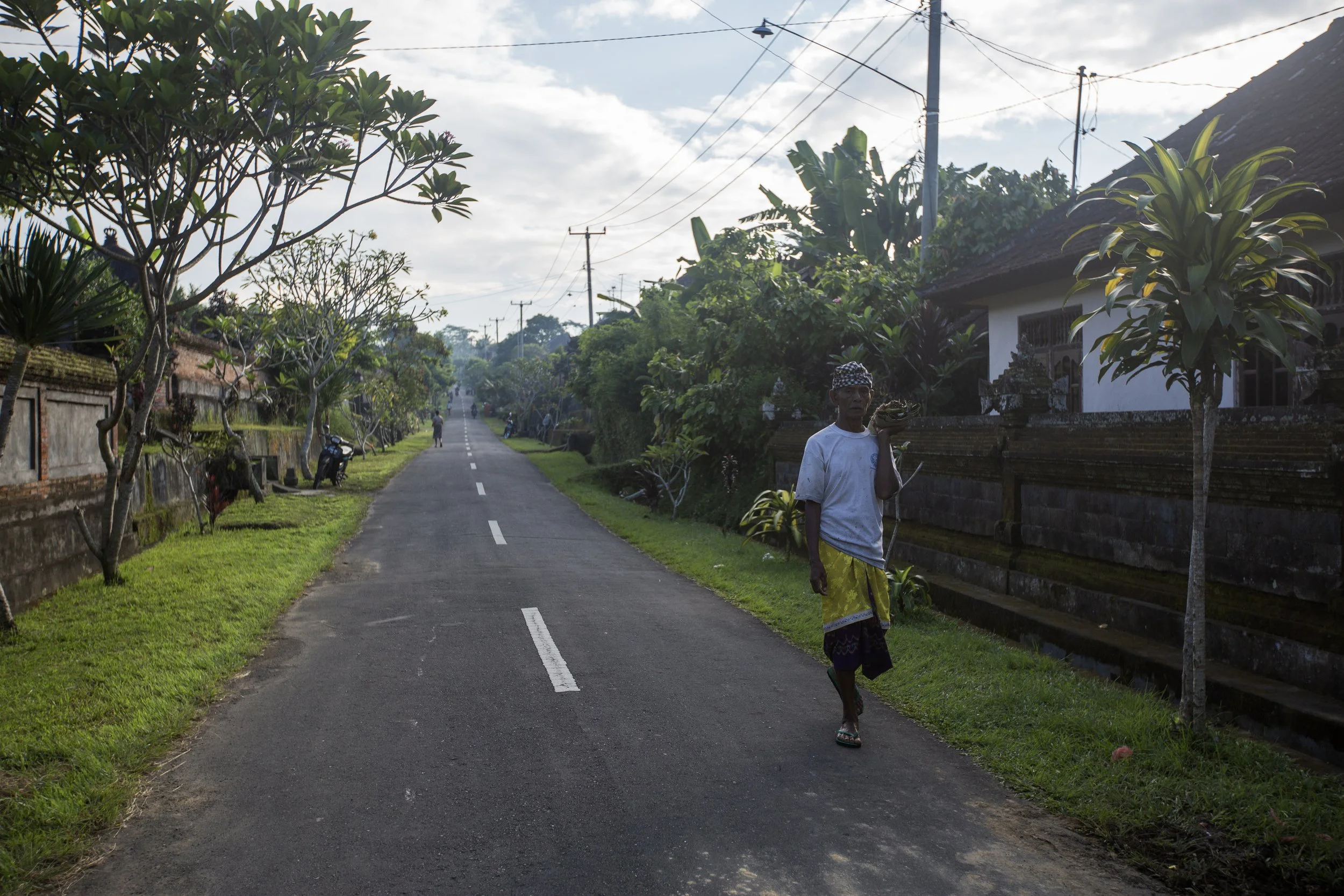 Un homme marche le long d'une route pavée dans un village tropical, portant une batte sur son épaule, entouré de végétation luxuriante et de maisons traditionnelles, sous un ciel partiellement nuageux.