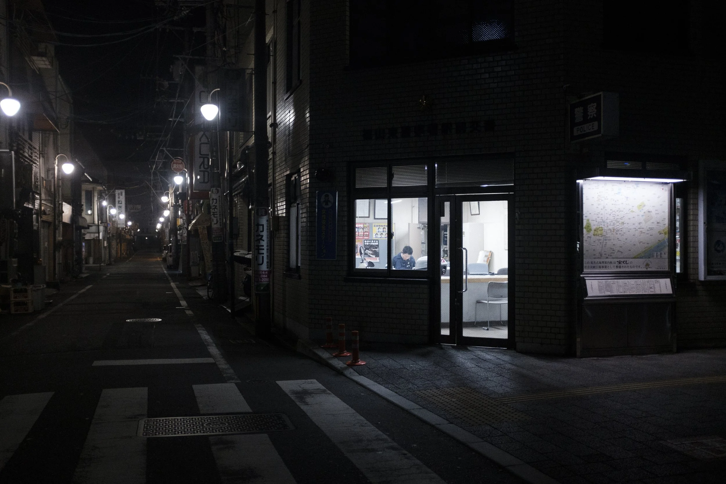 Une rue de nuit éclairée par des lampadaires, avec un bâtiment dans lequel on voit une personne assise à un bureau à travers une fenêtre, dans une ambiance calme et silencieuse.