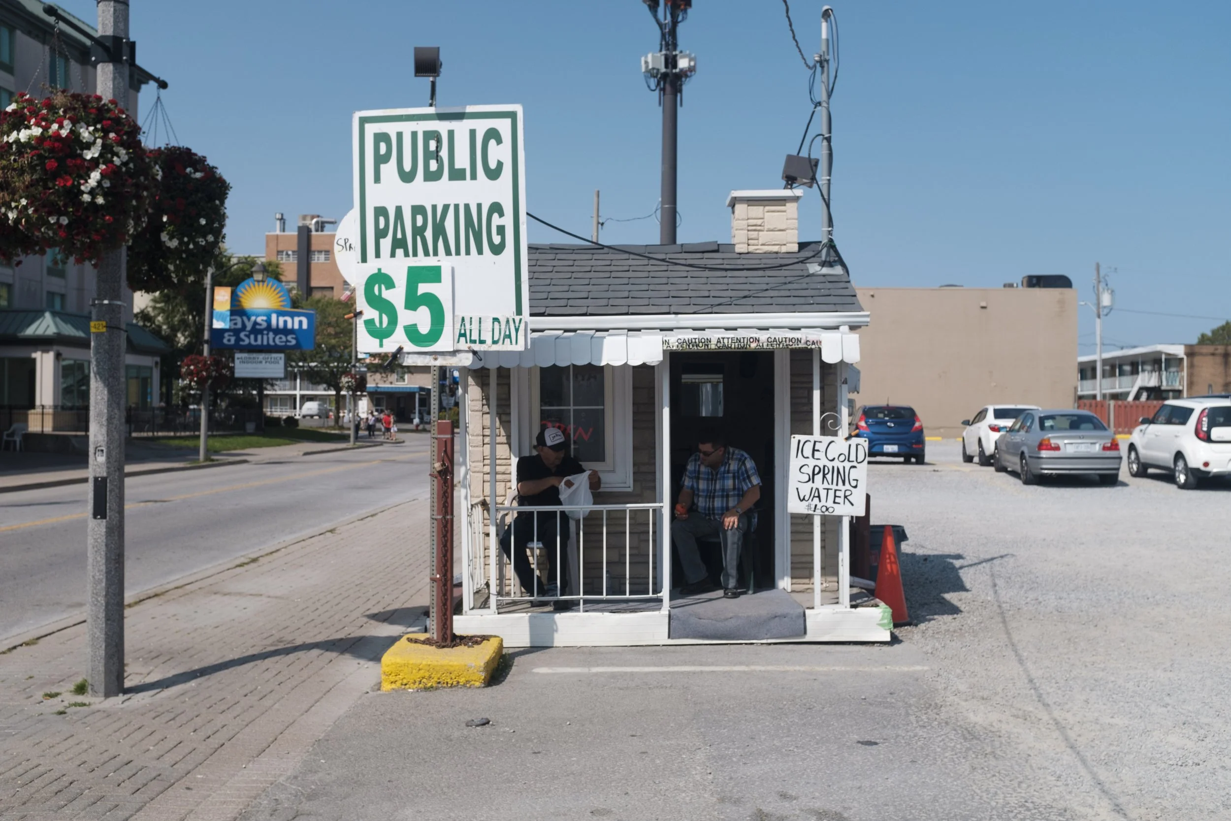 Petite cabine de vente d'eau avec deux personnes assises à l'intérieur et une pancarte indiquant "ICE COLD SPRING WATER $1.00" ainsi qu'une autre grande affiche précisant "PUBLIC PARKING $5 ALL DAY".
