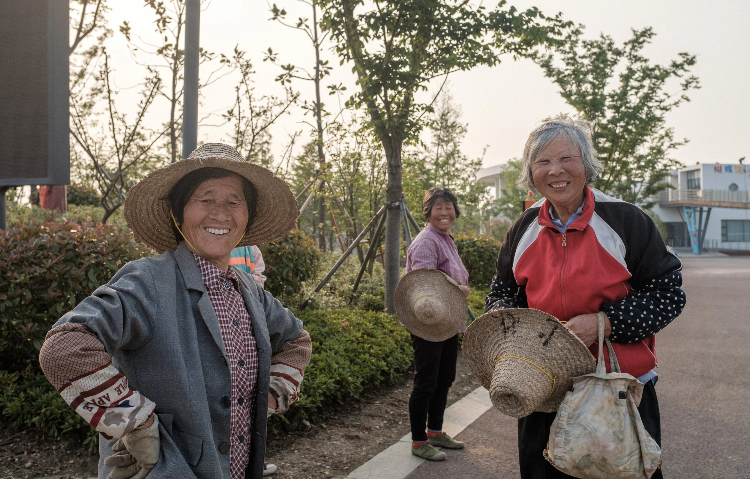 Quatre femmes souriantes portant des chapeaux en plein air, avec des arbres et un bâtiment moderne en arrière-plan.
