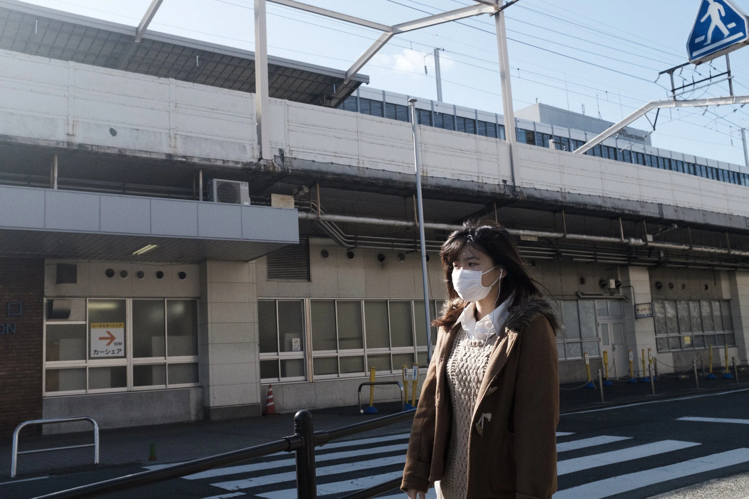 Une femme portant un masque protège le visage, vêtue d'un manteau marron et d'un pull beige, marchant à l'extérieur près d'une intersection de rue avec une structure de voie ferrée en arrière-plan.