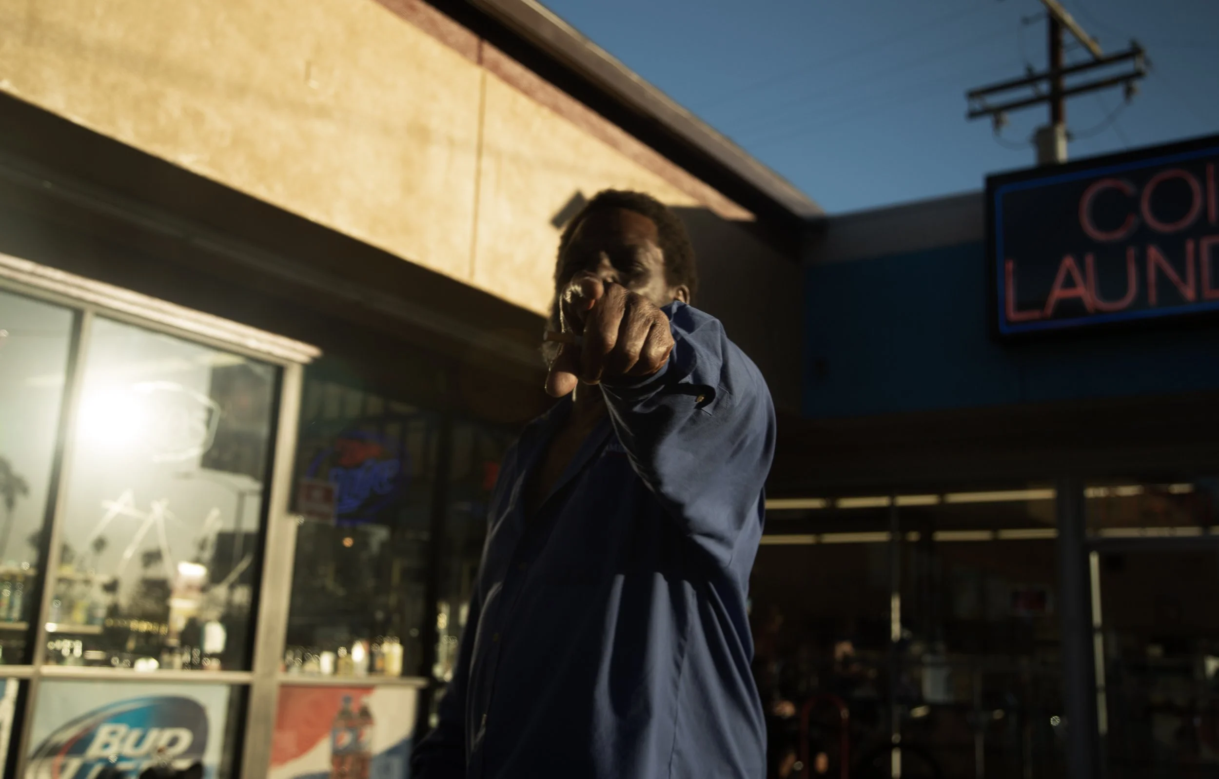 Un homme noir en chemise bleue pointe du doigt vers l'appareil photo devant un magasin, à l'extérieur au crépuscule