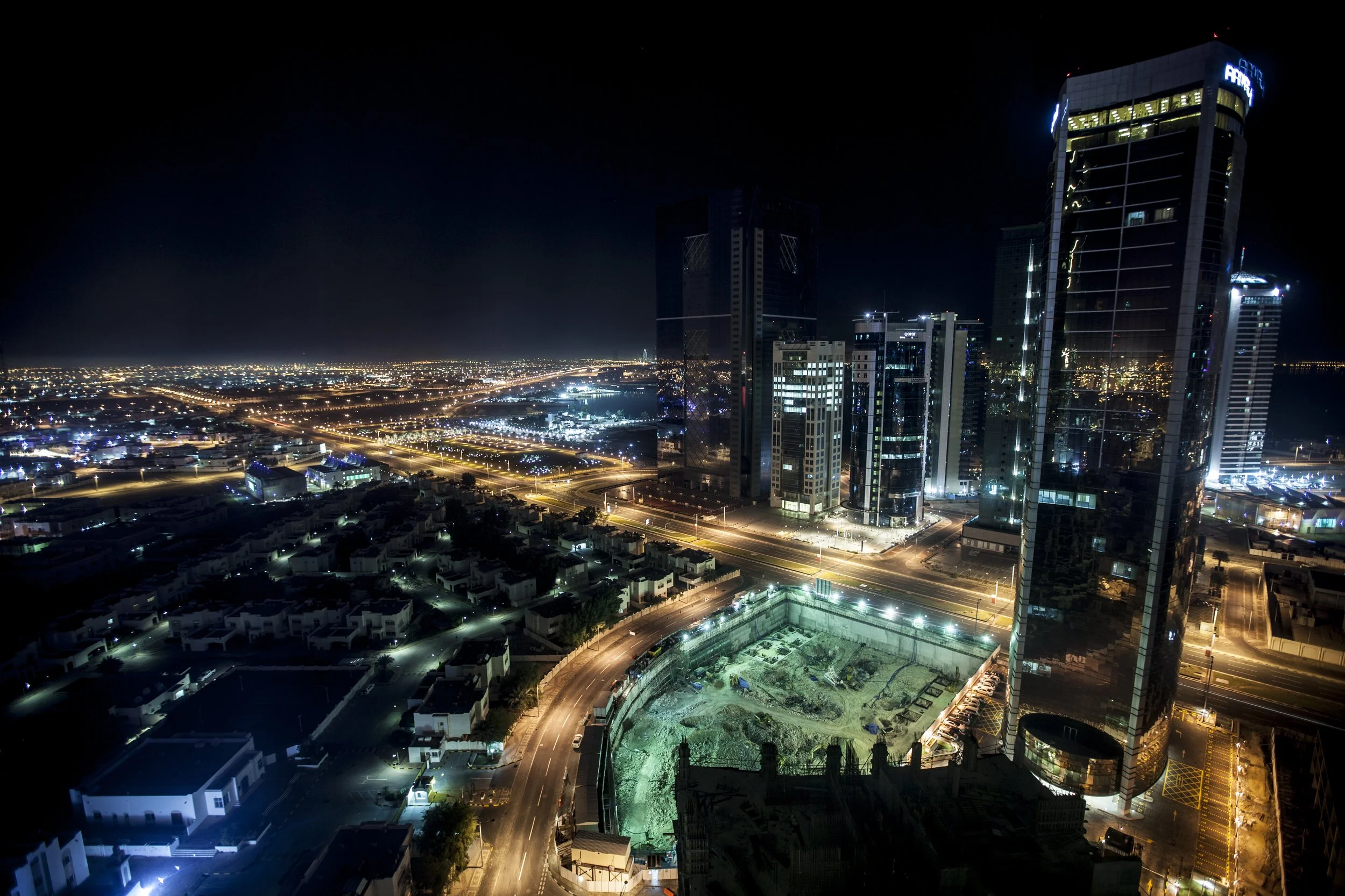 Cliché d'une ville moderne la nuit avec de grands immeubles en verre illuminés, des routes éclairées et un site de construction au premier plan.