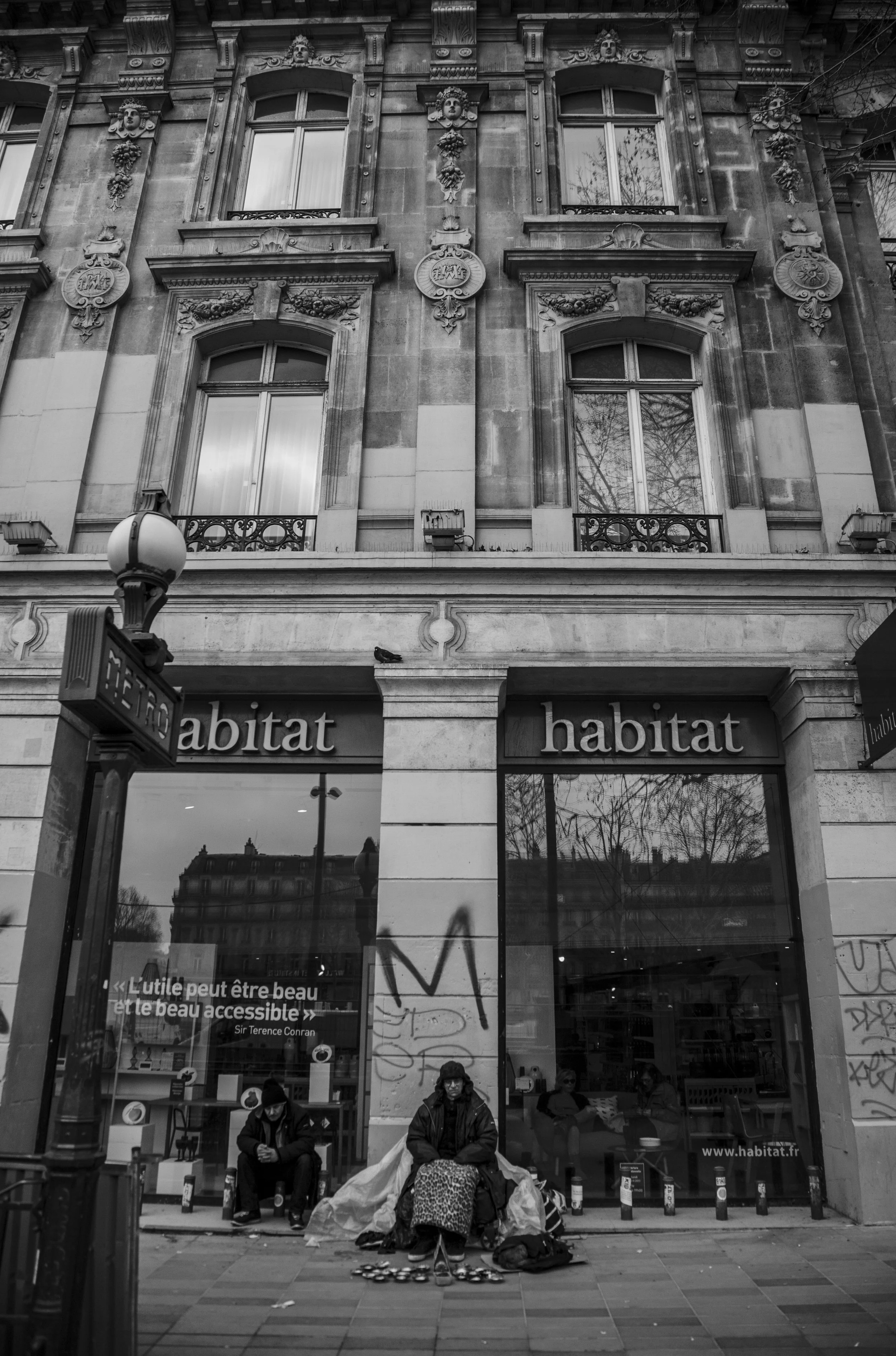 Photographie en noir et blanc de la façade d'un bâtiment Haussmannien avec des fenêtres ornées et des décorations classiques. Au rez-de-chaussée, un magasin Habitat avec deux vitrines et un message dans la fenêtre, et trois personnes assises devant, 