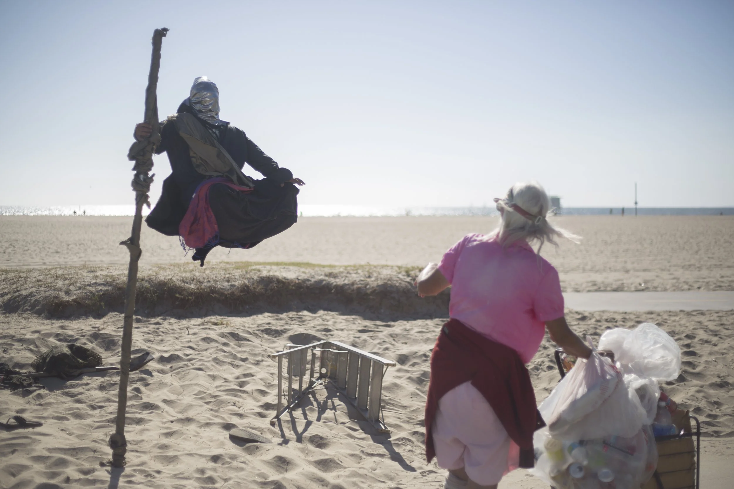 Une femme âgée portant un t-shirt rose, un t-shirt blanc en dessous, et une jupe rouge, ramasse des déchets et des bouteilles en plastique sur la plage, avec un coucher de soleil et la mer en arrière-plan.