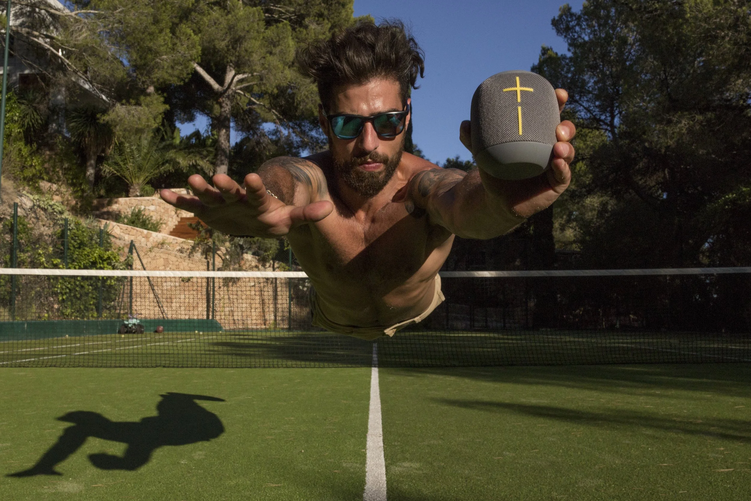 Un homme avec une barbe, portant des lunettes de soleil, est allongé en vol horizontal au-dessus d'un court de tennis, tenant une enceinte portable grise avec une croix jaune en face de la caméra, avec un fond de ciel bleu et d'arbres.