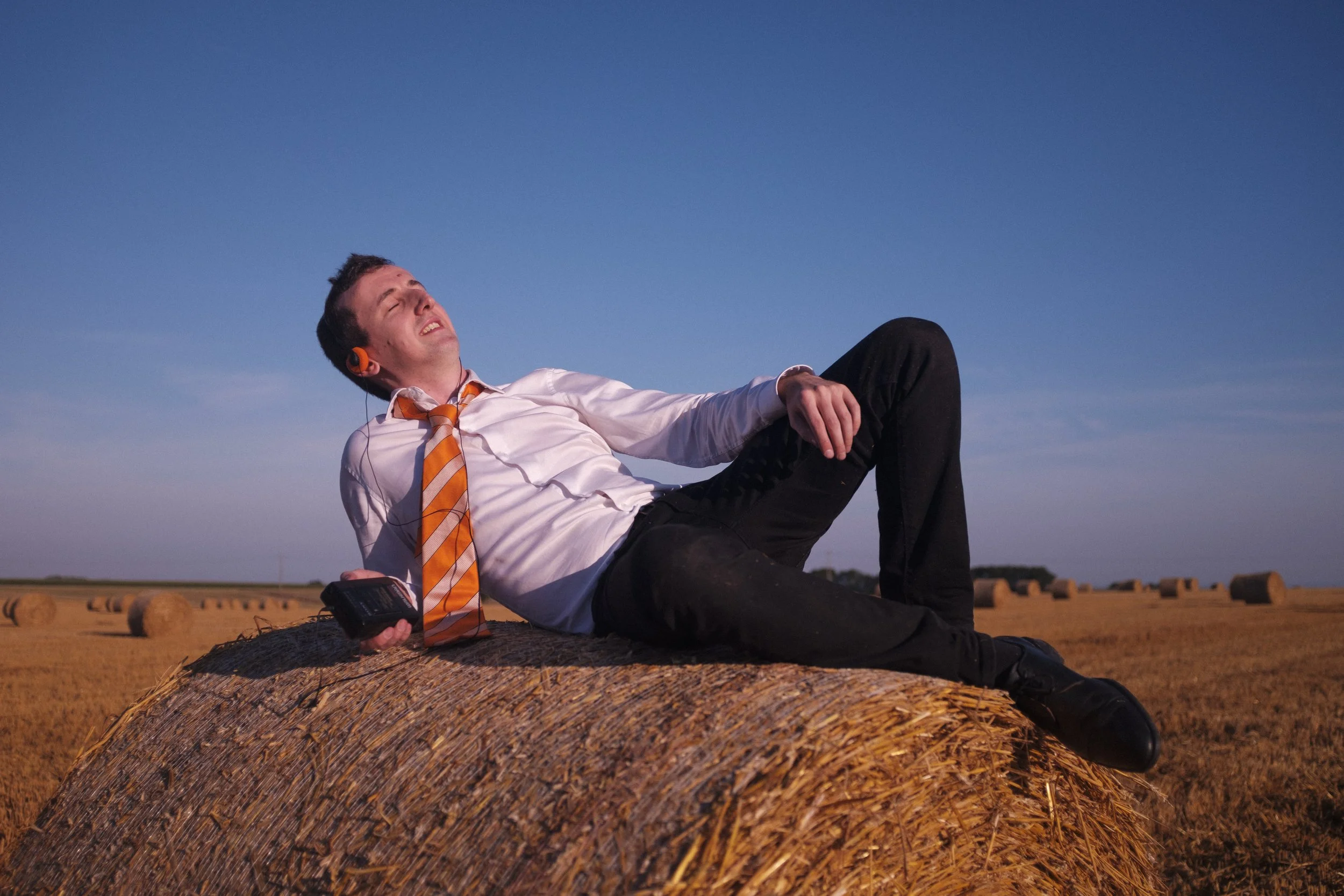 Un homme habillé en chemise blanche, cravate rayée orange et blanc, allongé sur une botte de foin dans un champ avec d'autres bottes en arrière-plan, sous un ciel dégagé, écoutant de la musique avec des écouteurs oranges, et tenant un appareil électr