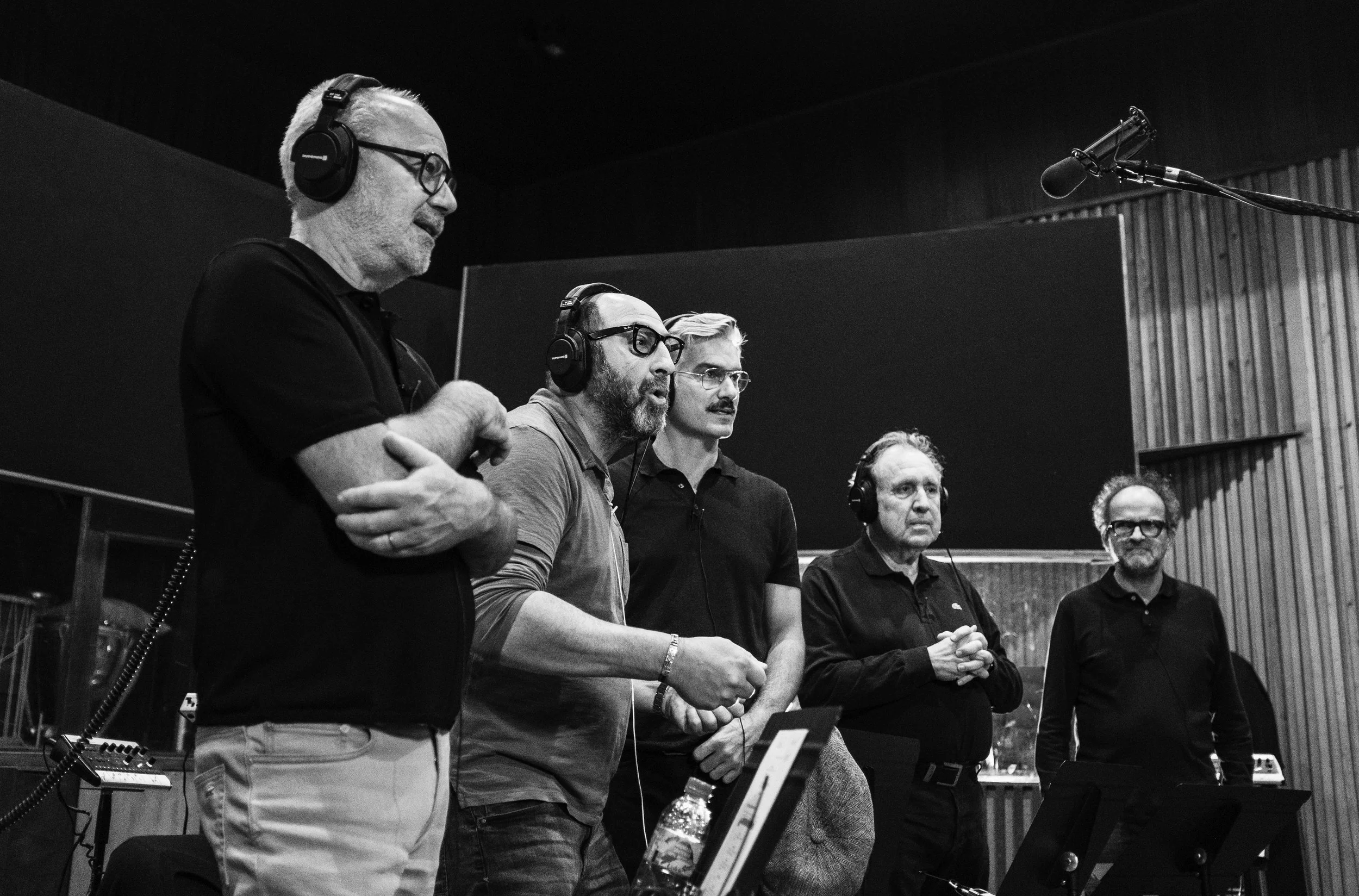 Cinq hommes avec casques d'écoute, enregistrement ou en studio, en train de parler ou d'écouter, dans une pièce avec murs en bois.